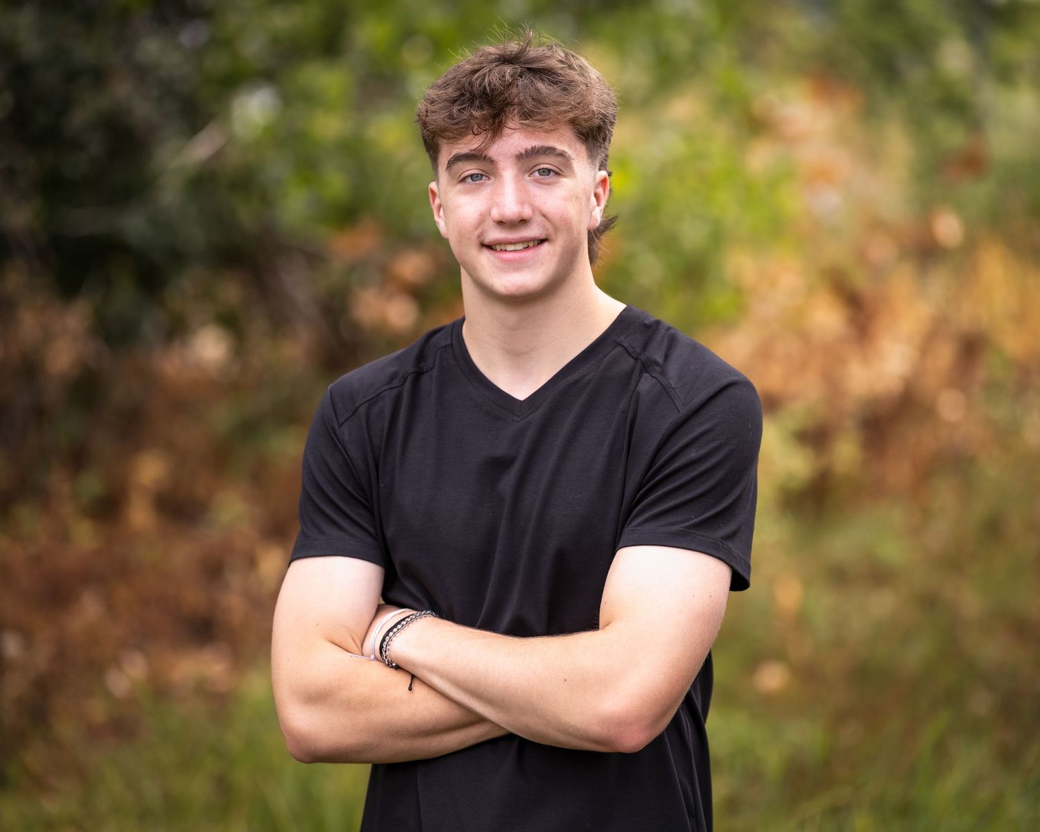 Smiling teen boy in black t-shirt with arms crossed standing in front of autumn foliage.