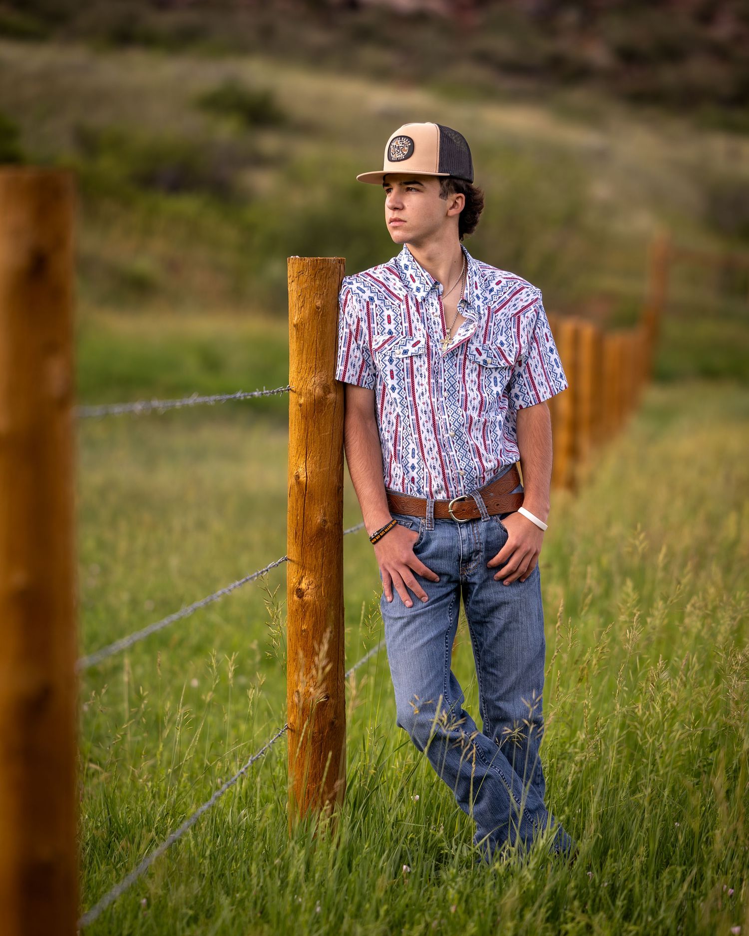 Young man in western shirt and trucker hat leaning on fence post in green field.