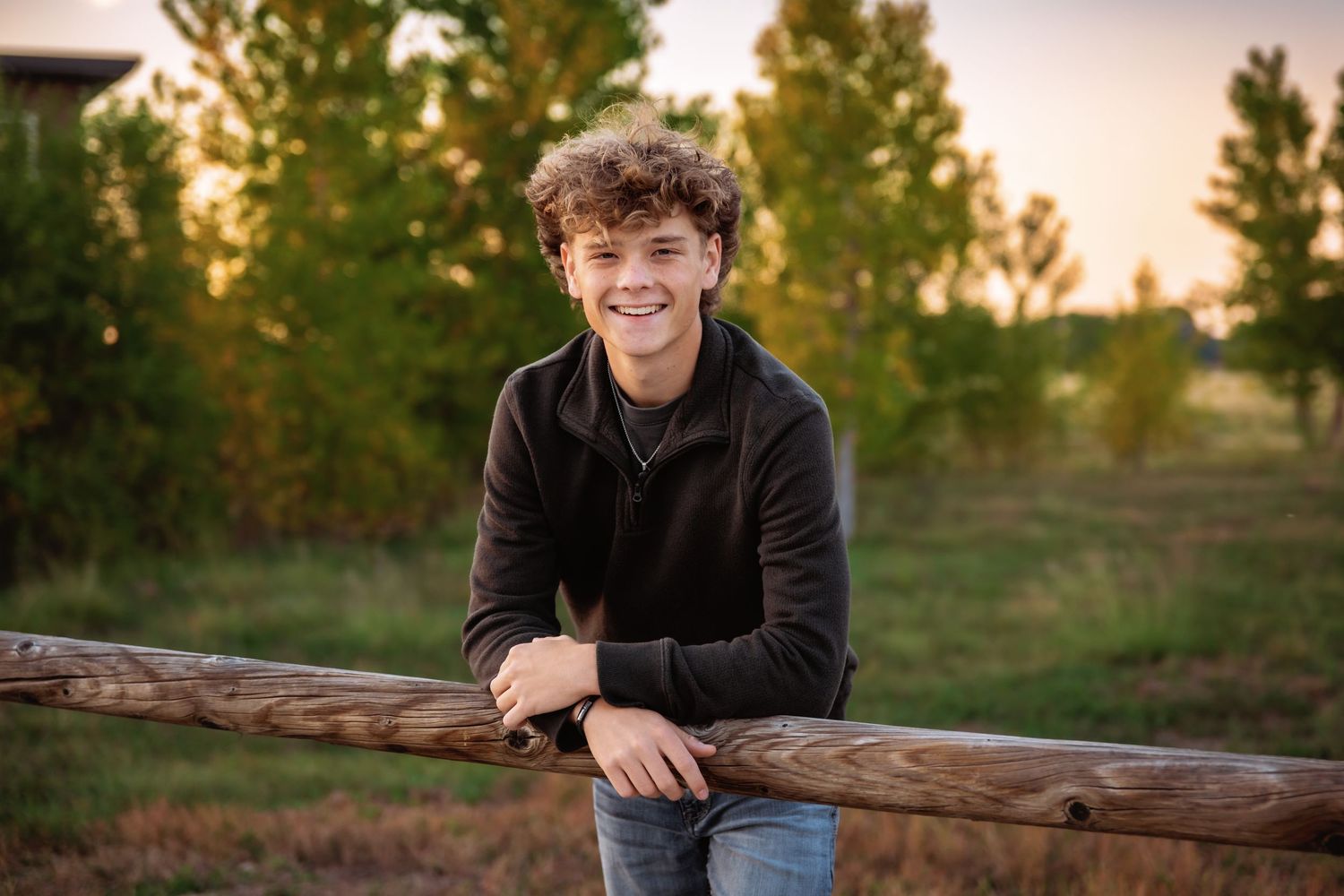 Smiling teenage boy with curly hair leaning on a wooden fence rail in a sunny outdoor field.