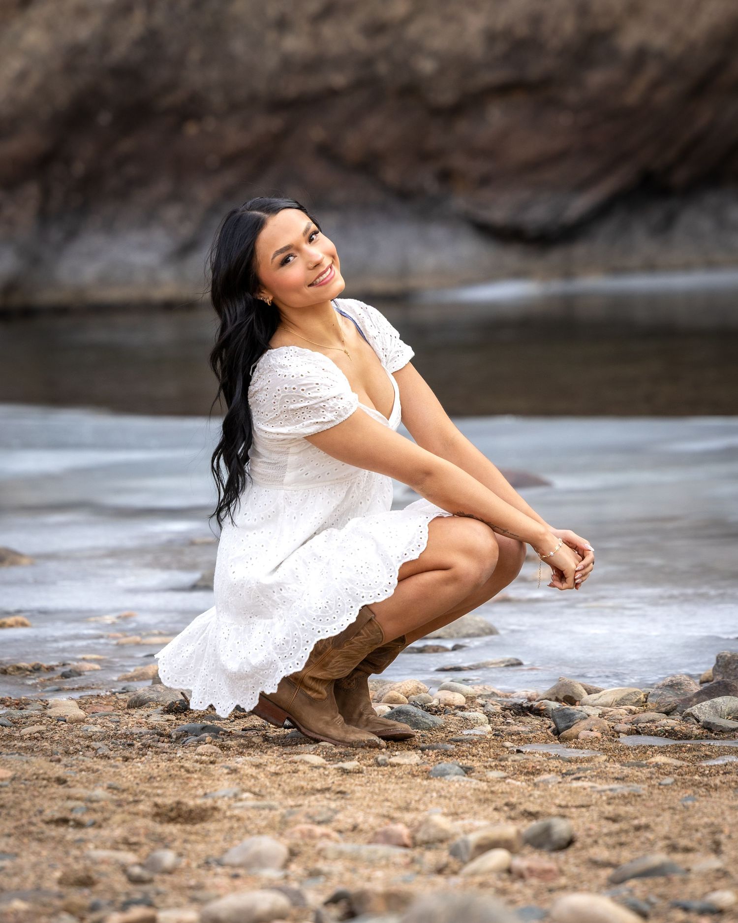 Young woman in a white dress and brown boots crouching by a rocky riverside smiling over her shoulder.