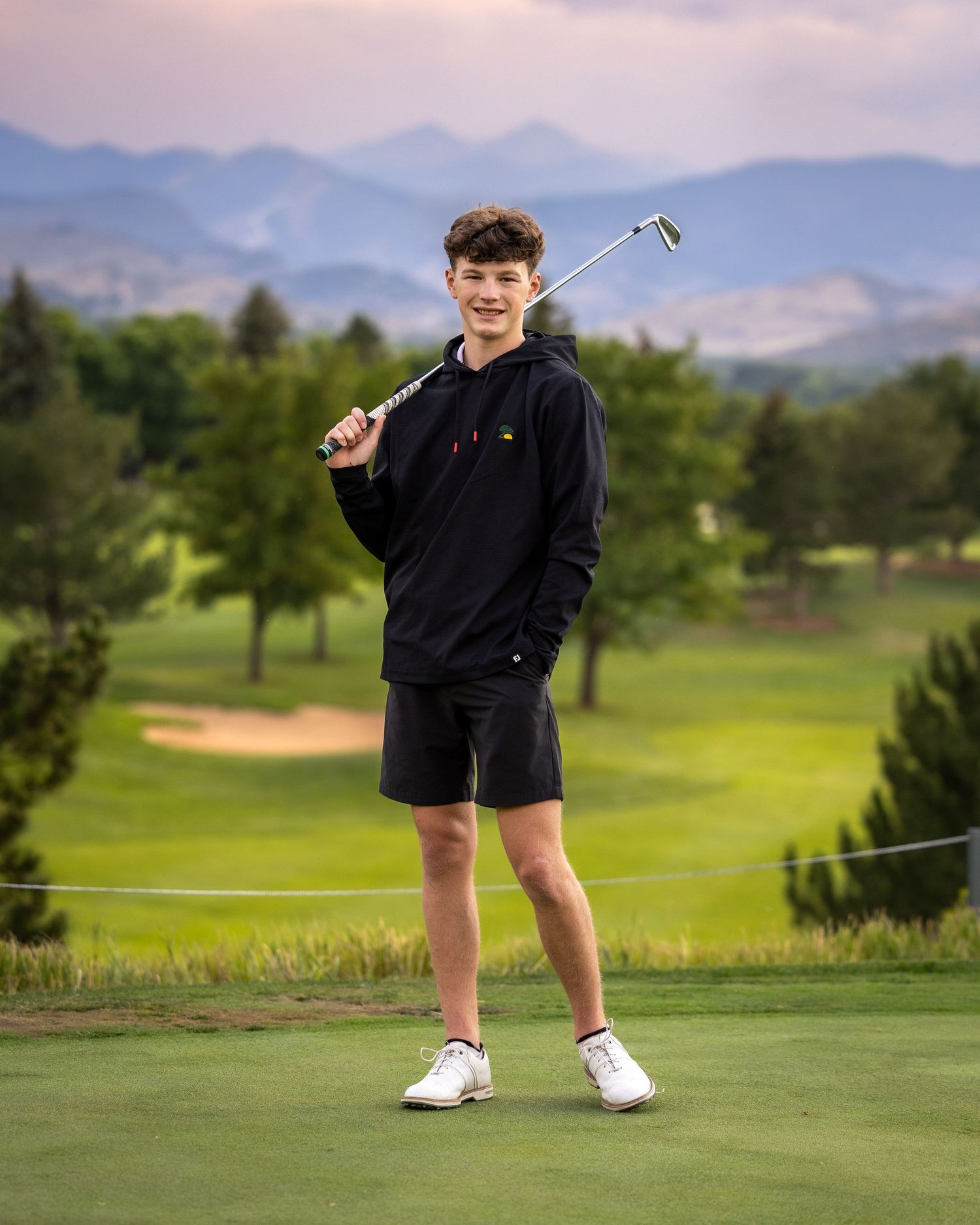 Teen boy in black hoodie poses with golf club on lush green golf course with mountain views.