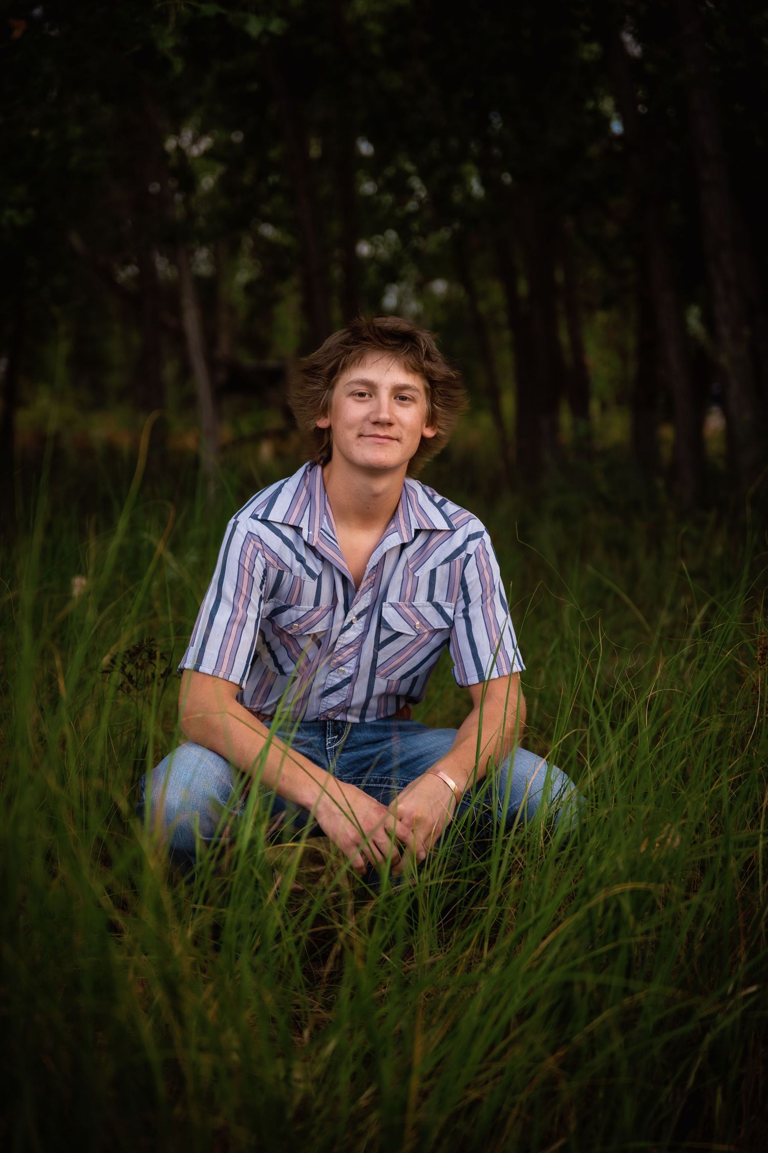 Smiling teen boy in striped western shirt sitting cross-legged in tall green grass.