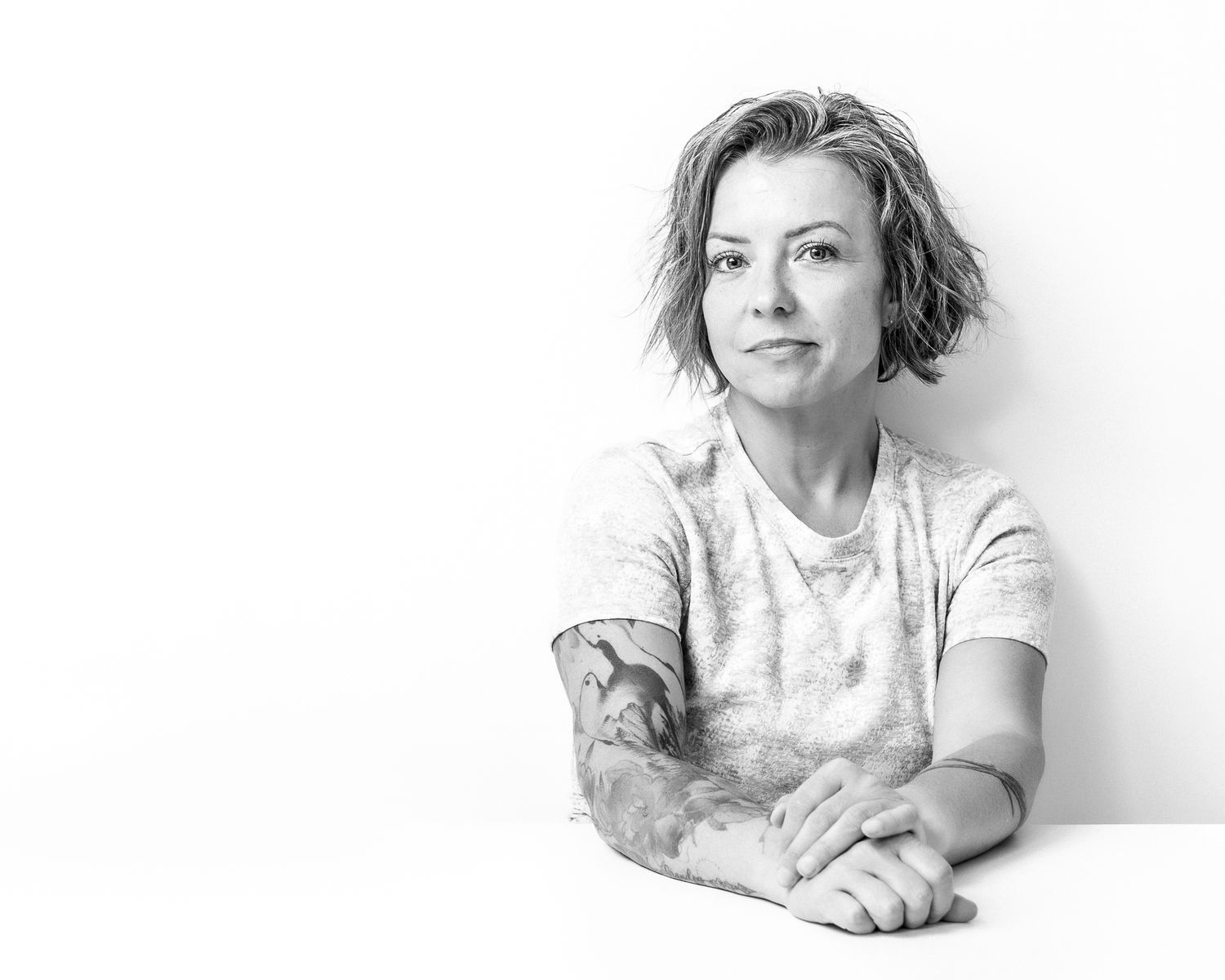 Black and white portrait of a woman with short hair and tattoos sitting casually at a table.