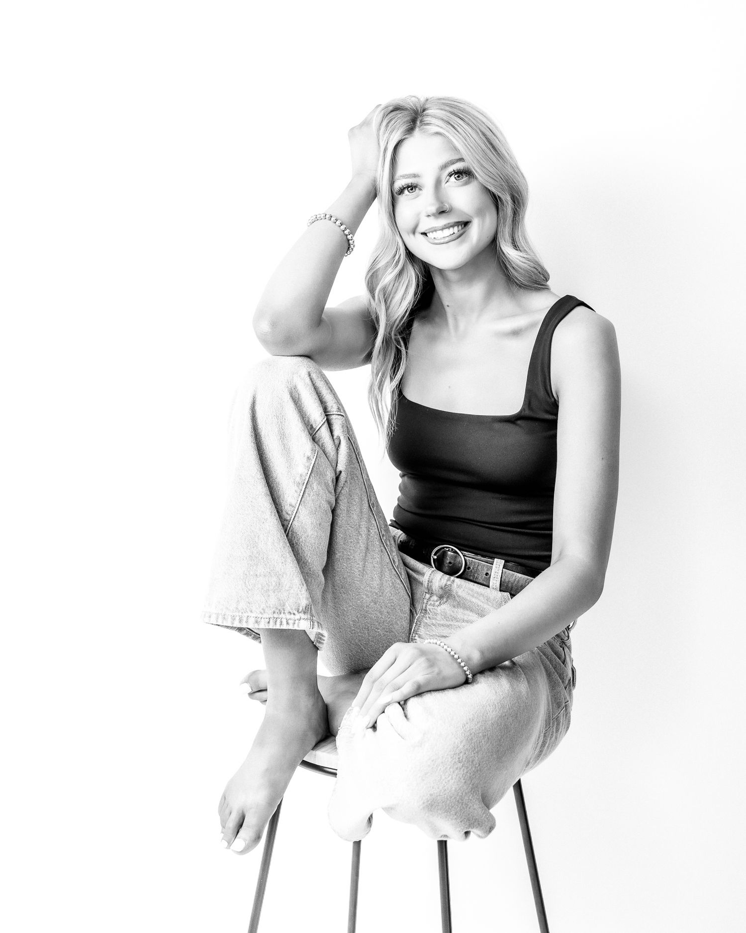 Black and white photo of a smiling blonde woman sitting on a stool in jeans and tank top.