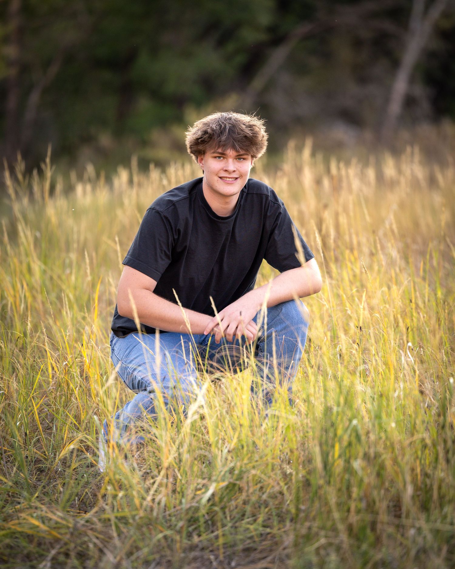 Smiling teen boy in black t-shirt crouches in tall golden grass field at golden hour.