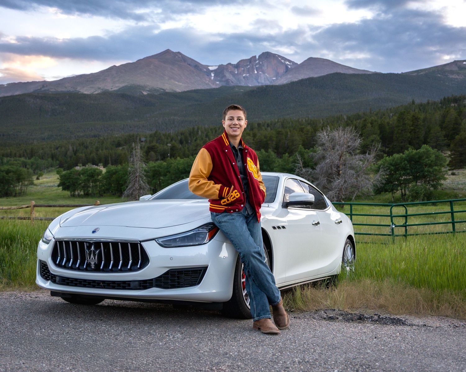 Young man in varsity jacket leans against white Maserati with Rocky Mountain backdrop at sunset.