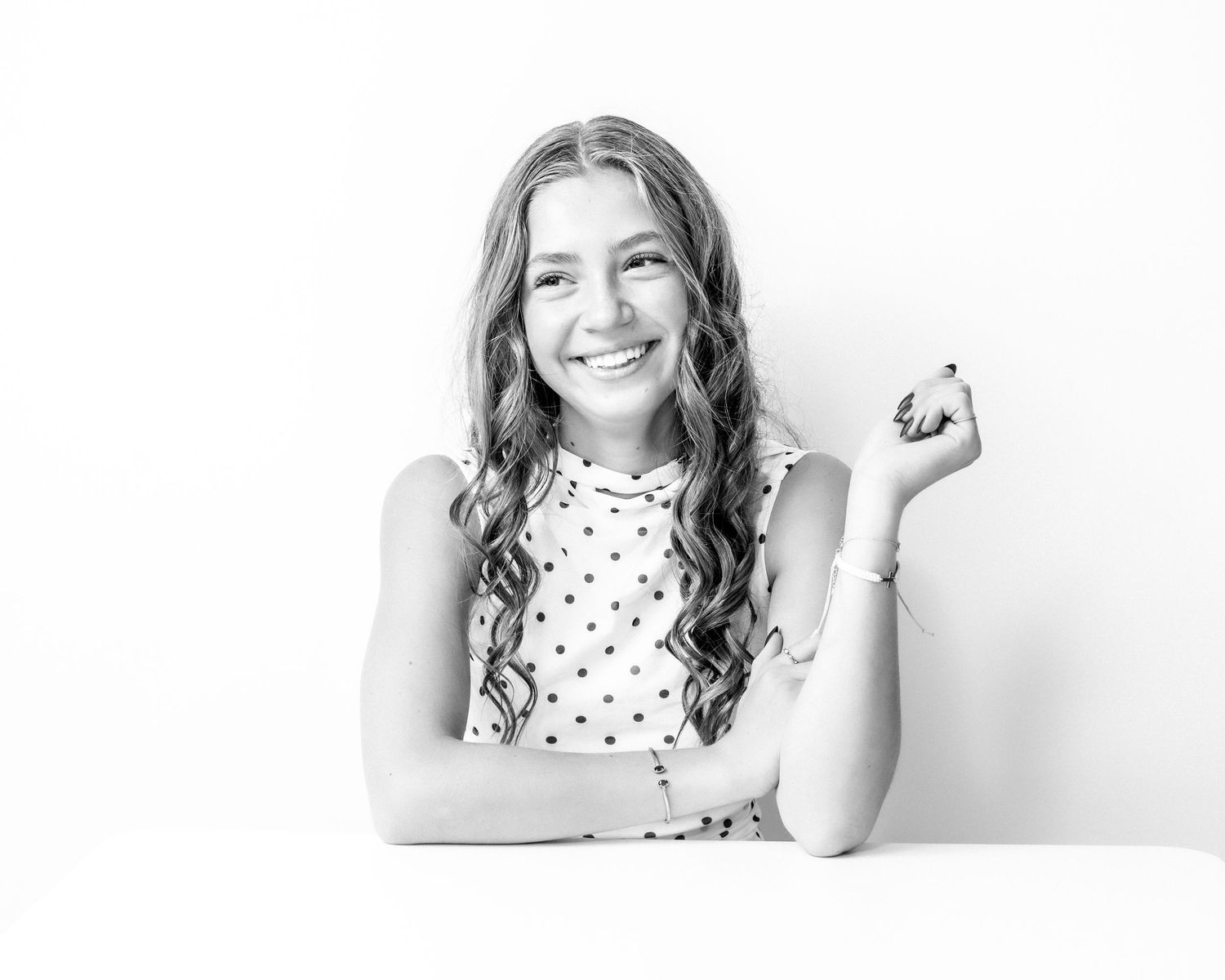 Smiling young woman with curly hair poses at a table in a polka dot top in a black and white photo.