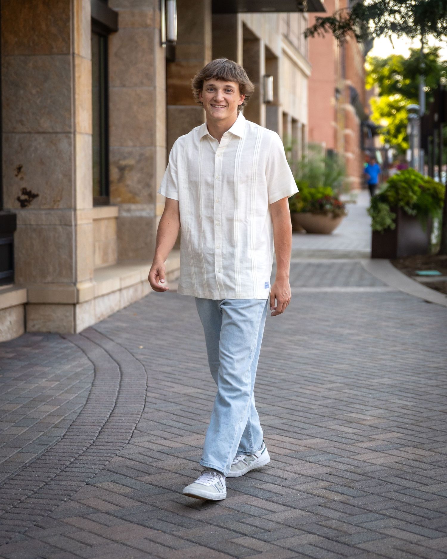Teenage boy in a white shirt and light jeans walking on a brick sidewalk in an urban setting.