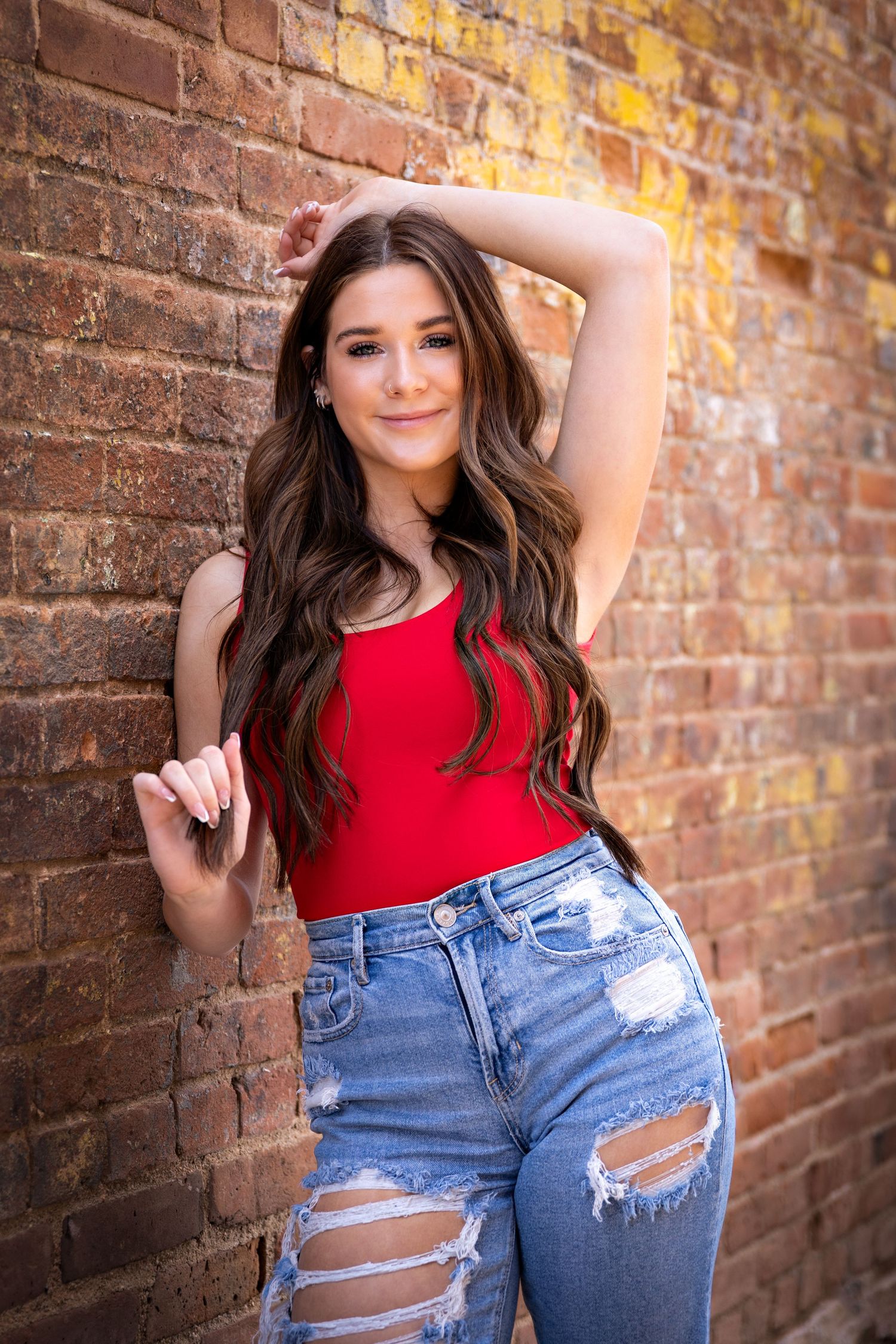 Brunette girl in red top and ripped jeans posing against rustic brick wall.