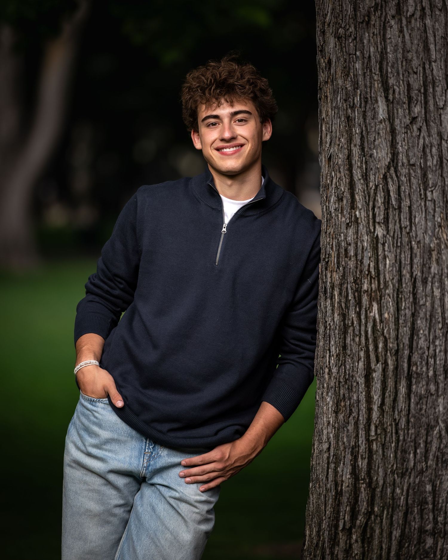 Smiling teen with curly hair leans against tree trunk wearing navy quarter-zip pullover outdoors.