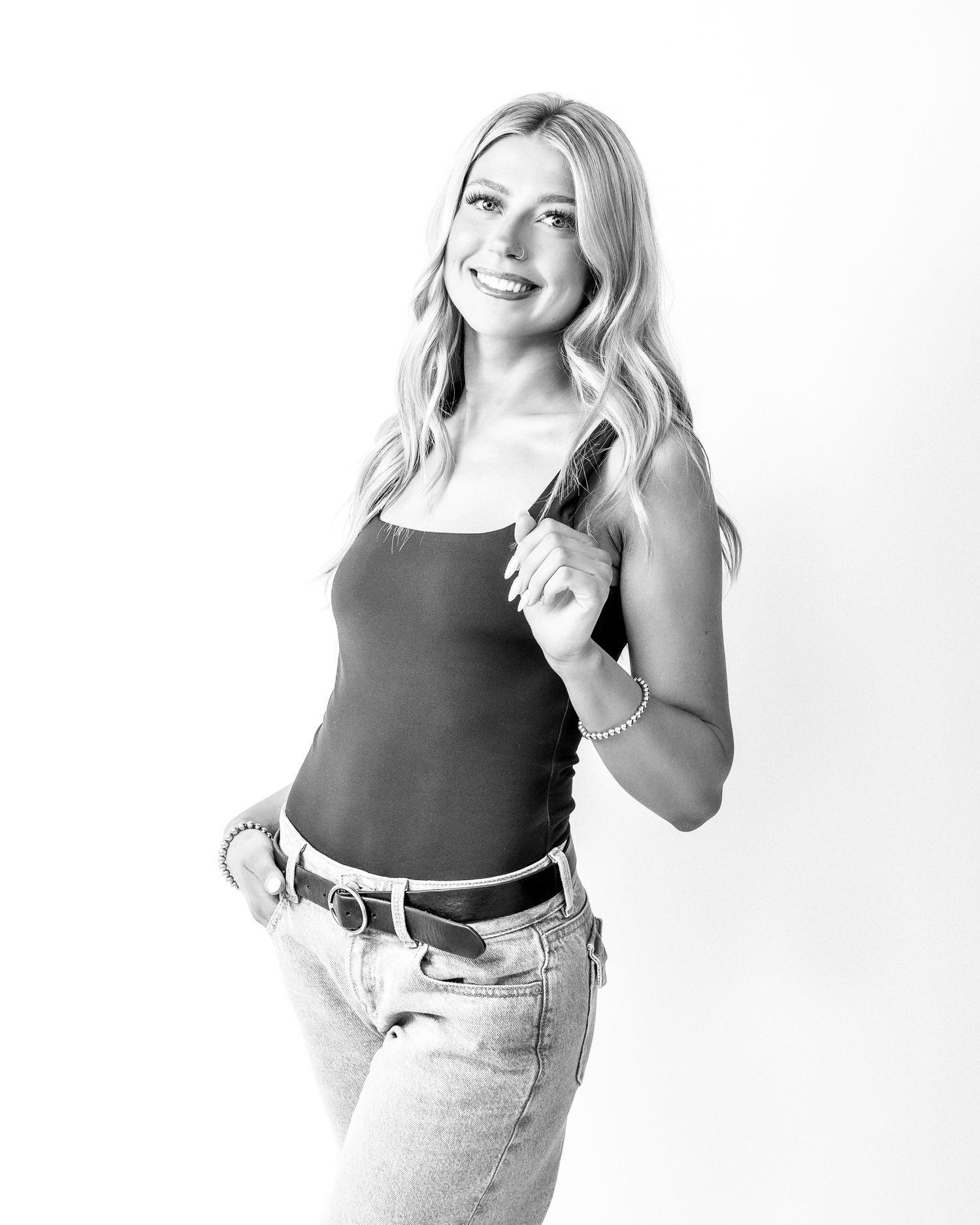 Black and white photo of smiling blonde woman in tank top and jeans holding a handgun in a studio setting.