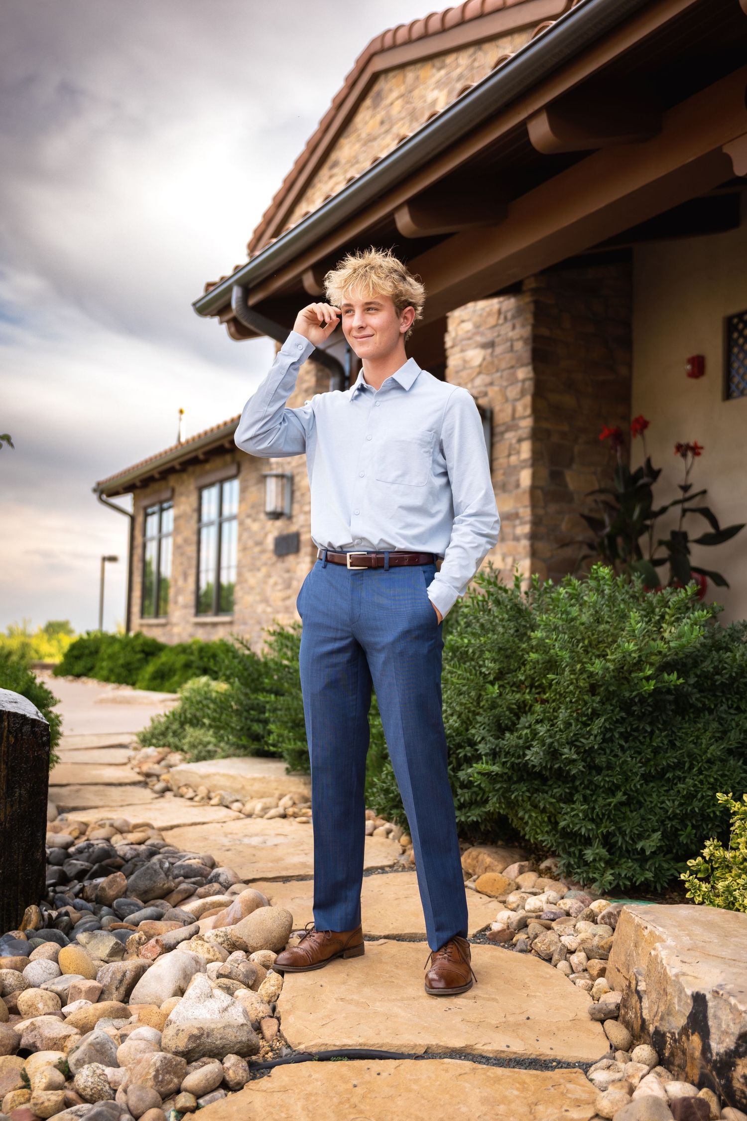 Stylish young blond man in blue dress pants and button-up shirt posing outside a stone building.
