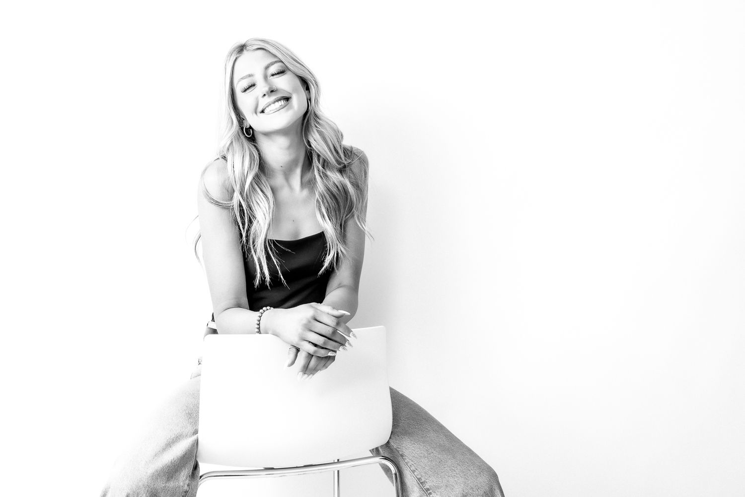 Smiling blonde woman sitting relaxed in black top, black and white portrait photography studio shot.