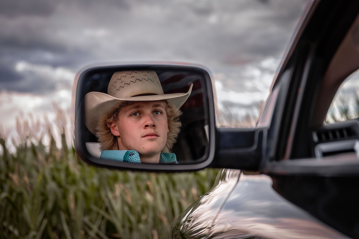 Young man in cowboy hat reflected in truck side mirror with stormy sky background.