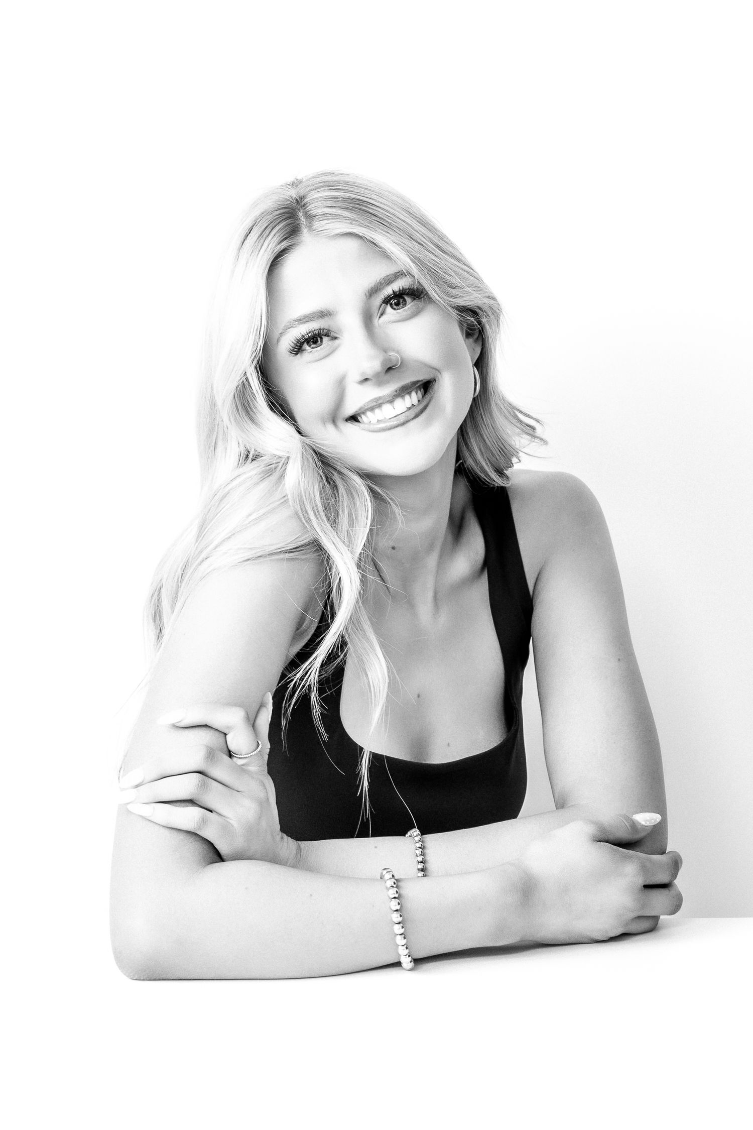 Black and white portrait of a smiling blonde woman in a black top, wearing a bracelet, posing at a table.