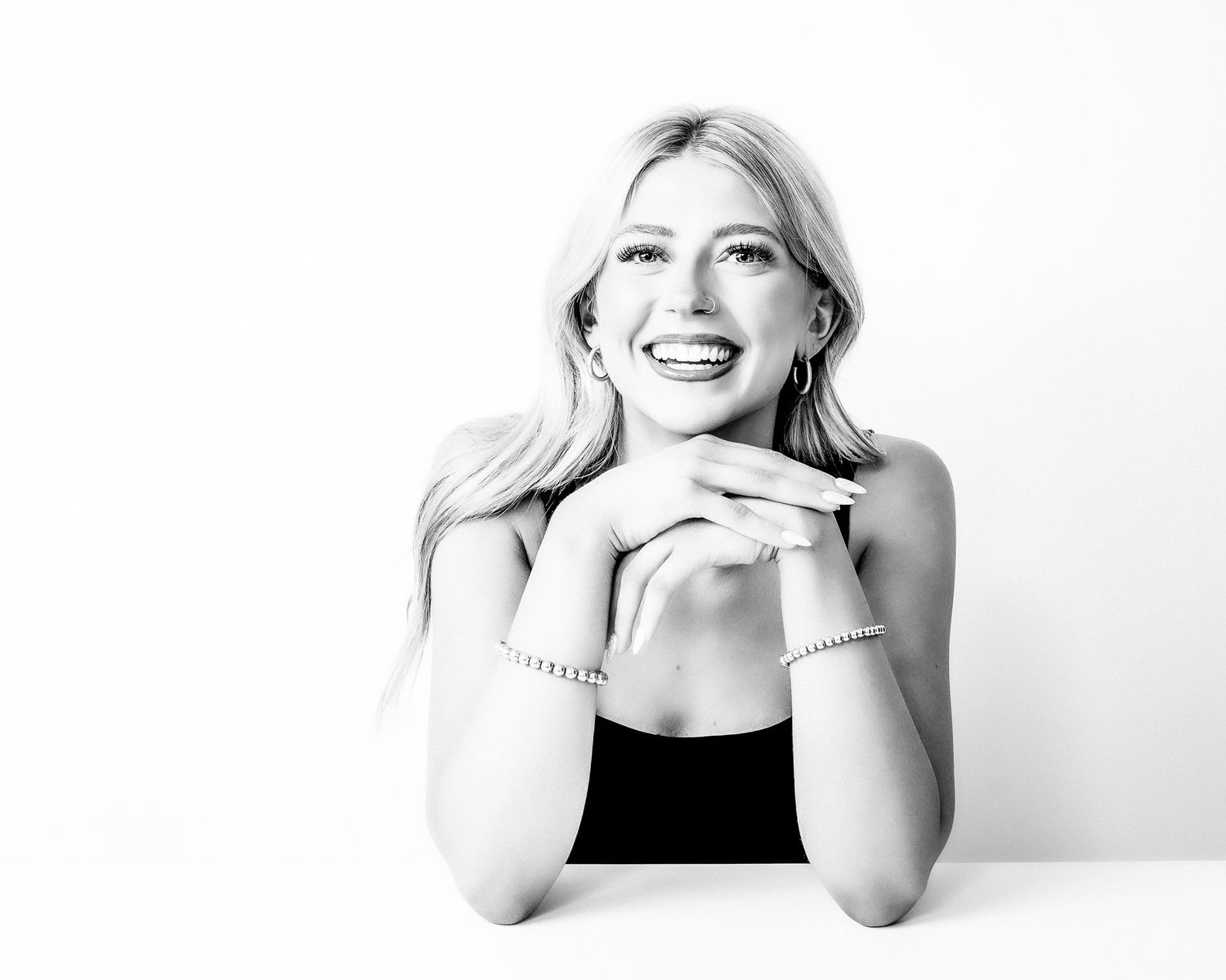 Smiling blonde woman posing with hands clasped under chin, wearing black top and bracelets, black and white photo.