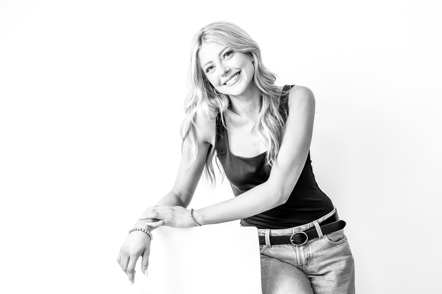 Smiling blonde woman in black tank top and jeans leaning on white cube in black and white studio photo.