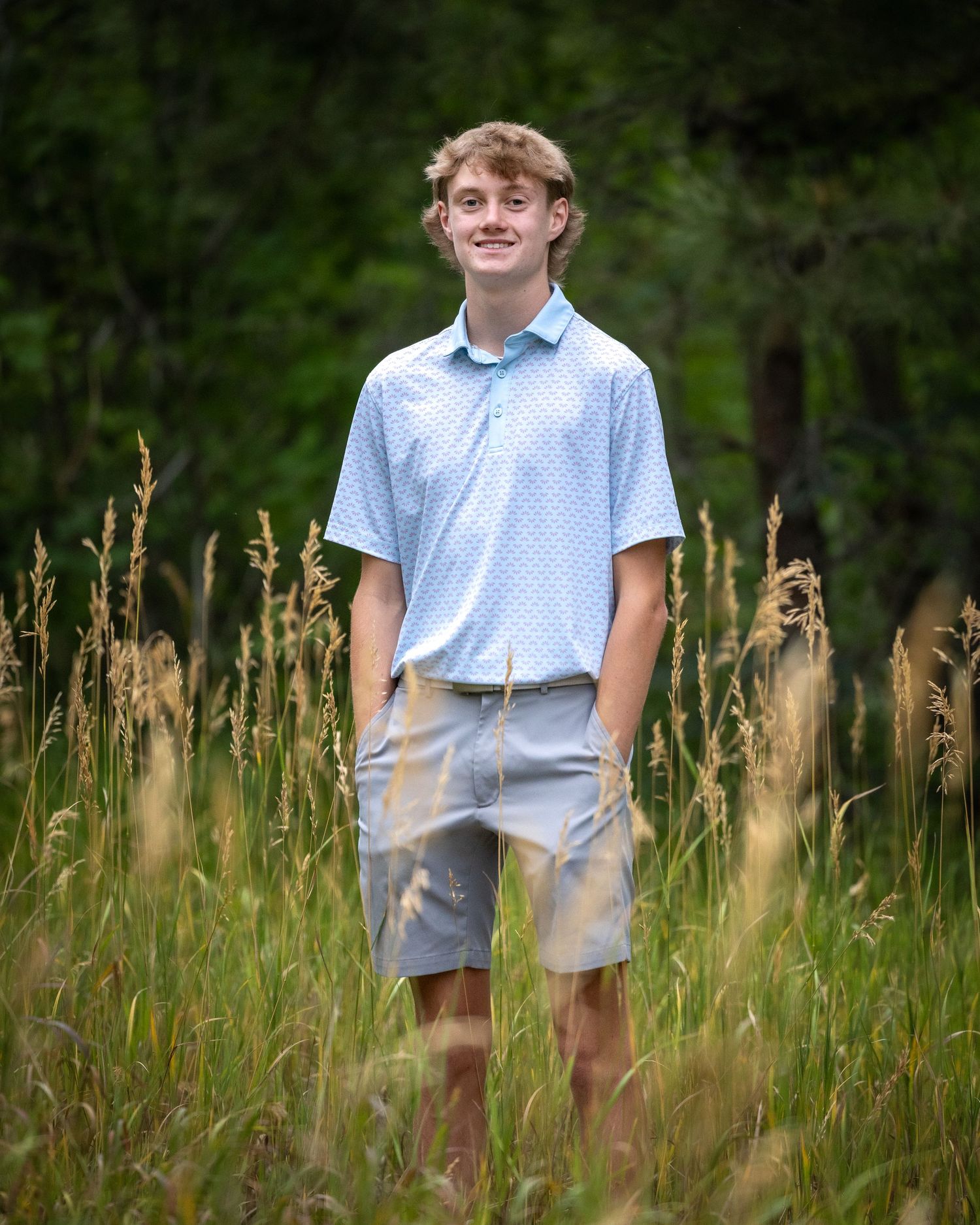 Smiling teen boy in light blue polo stands in tall grass field surrounded by trees.