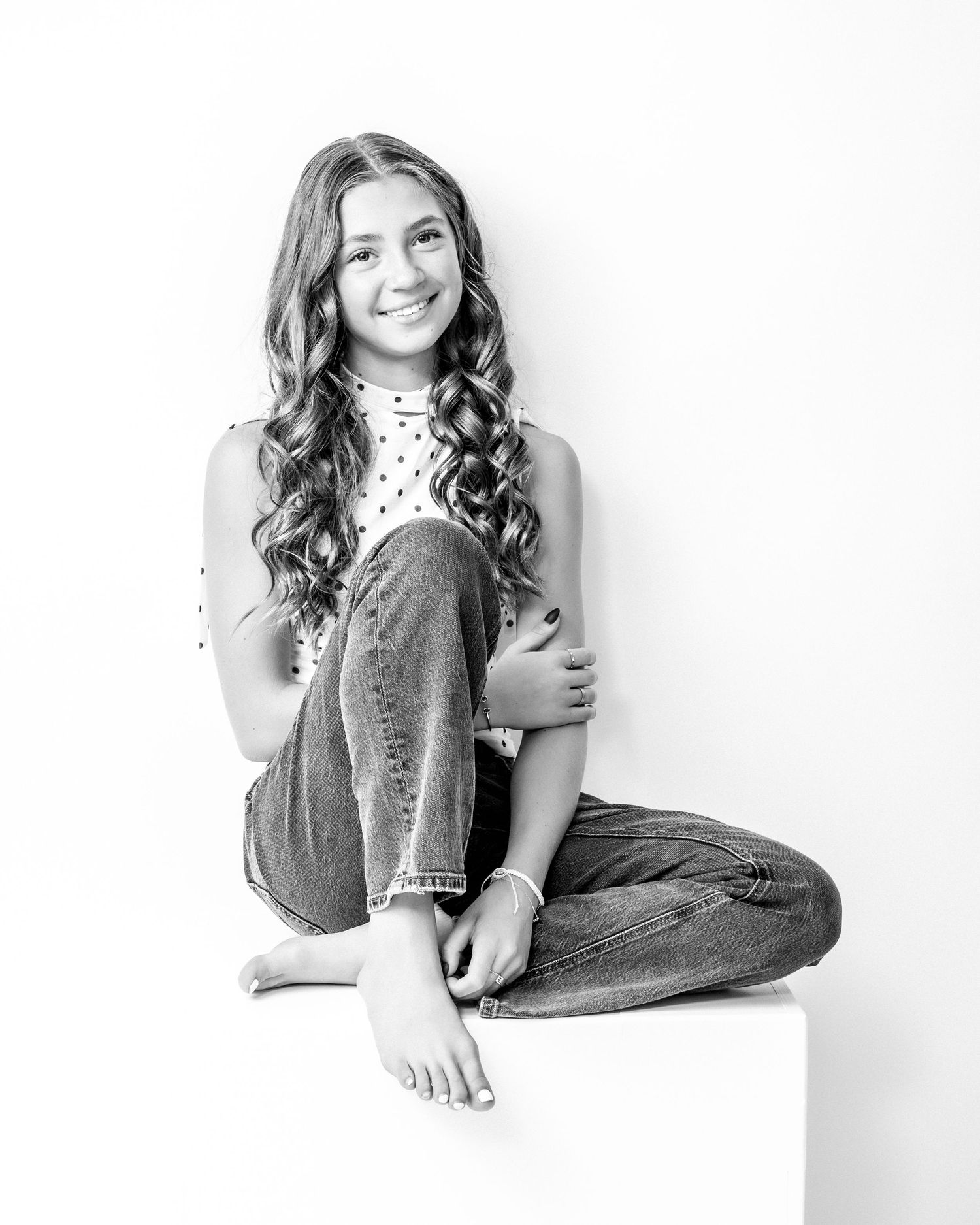 Black and white photo of a smiling teen girl with curly hair sitting cross-legged on a cube.