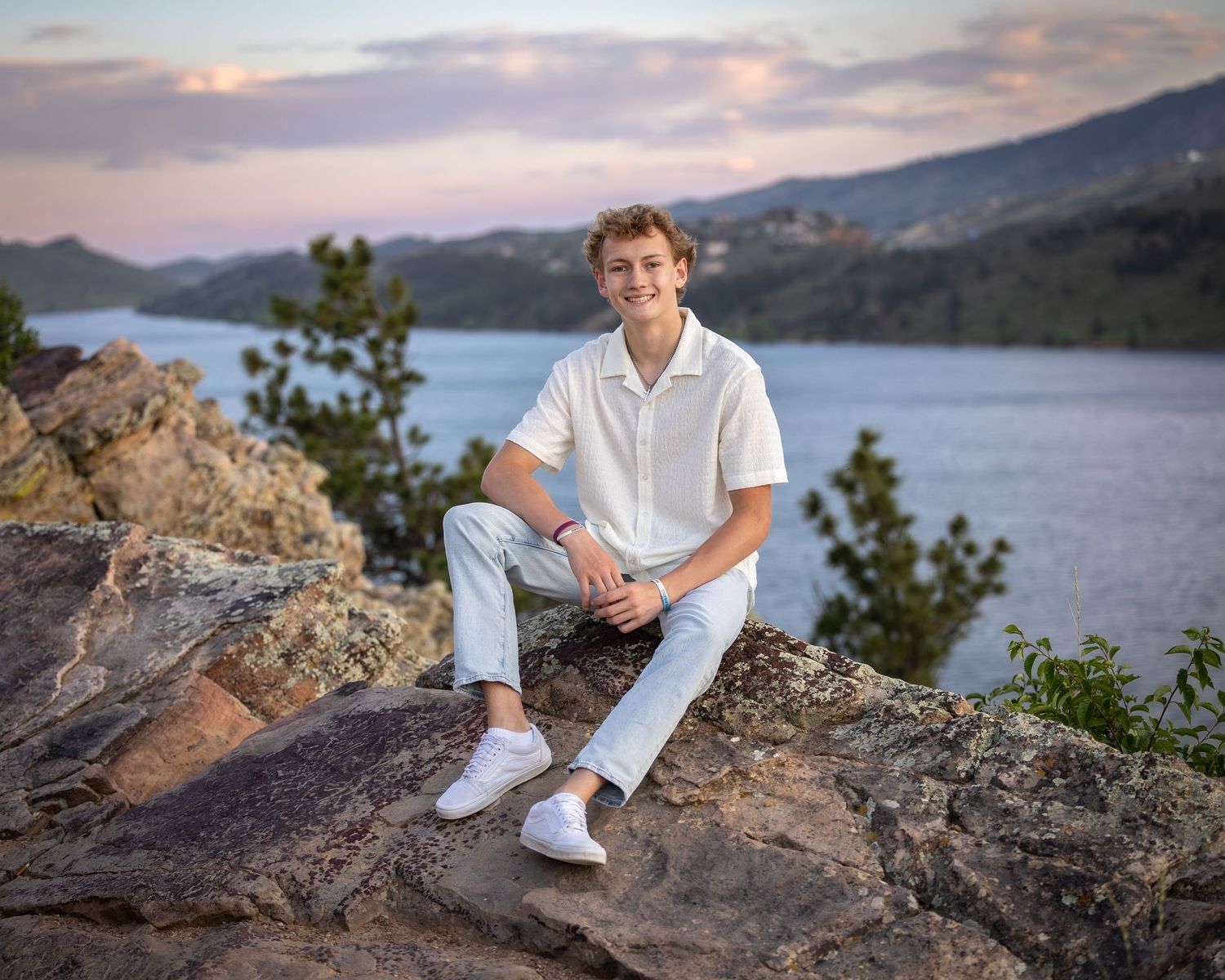 Young man in white shirt sits on rocky overlook above scenic mountain lake at sunset.