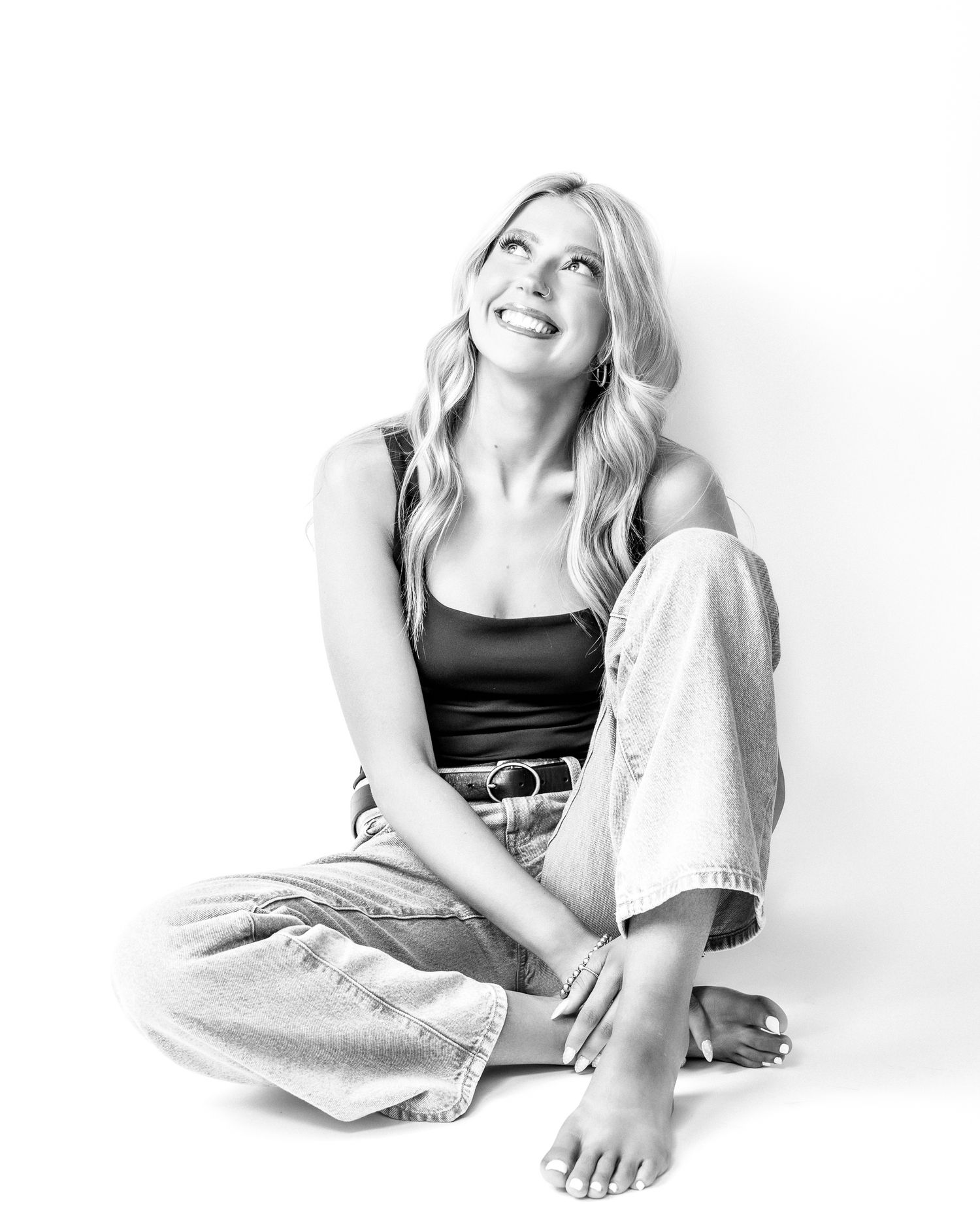 Black and white photo of a smiling blonde woman sitting cross-legged on the floor in jeans and a tank top.