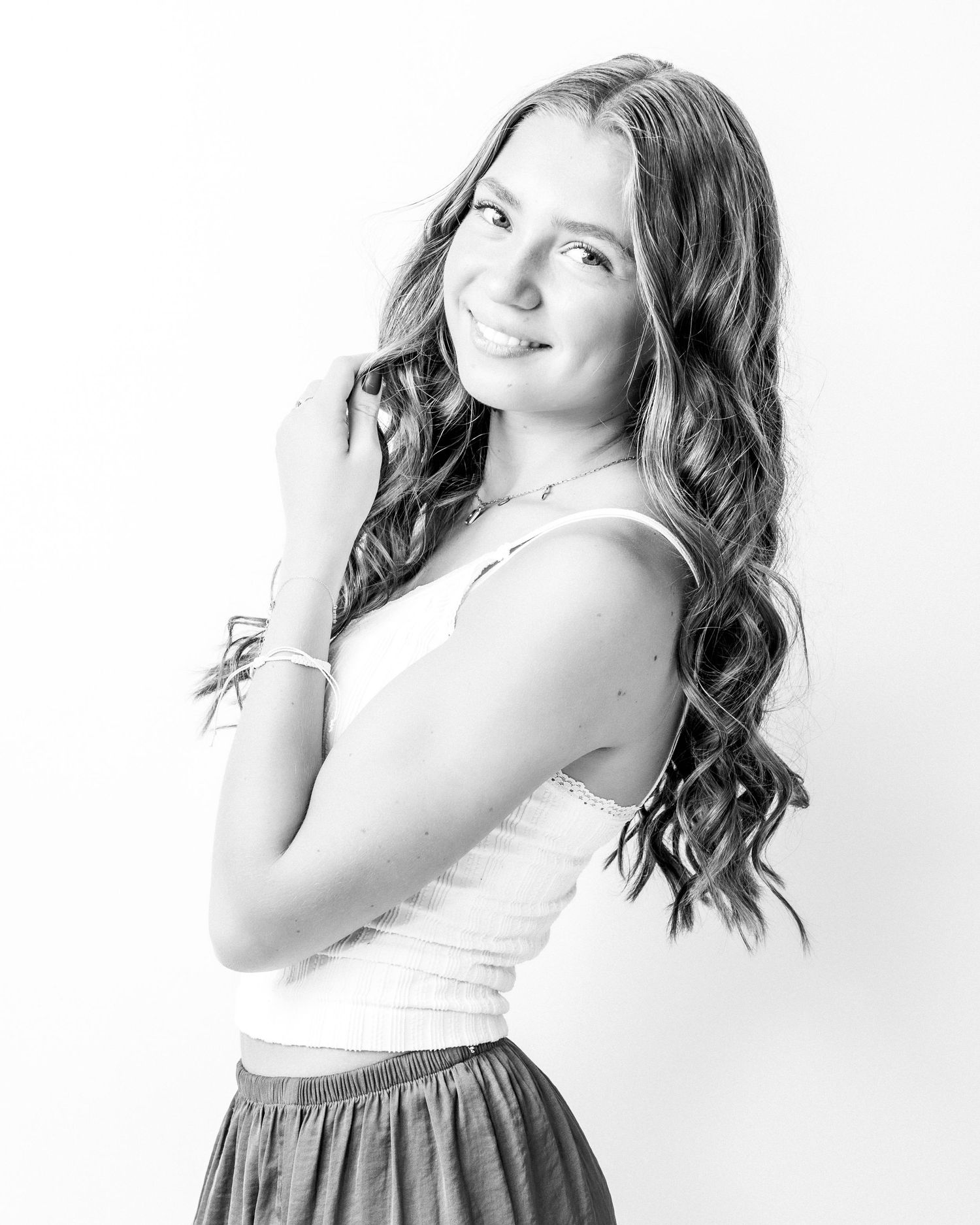 Young woman with long wavy hair smiles over her shoulder in a white tank top in a black and white portrait.