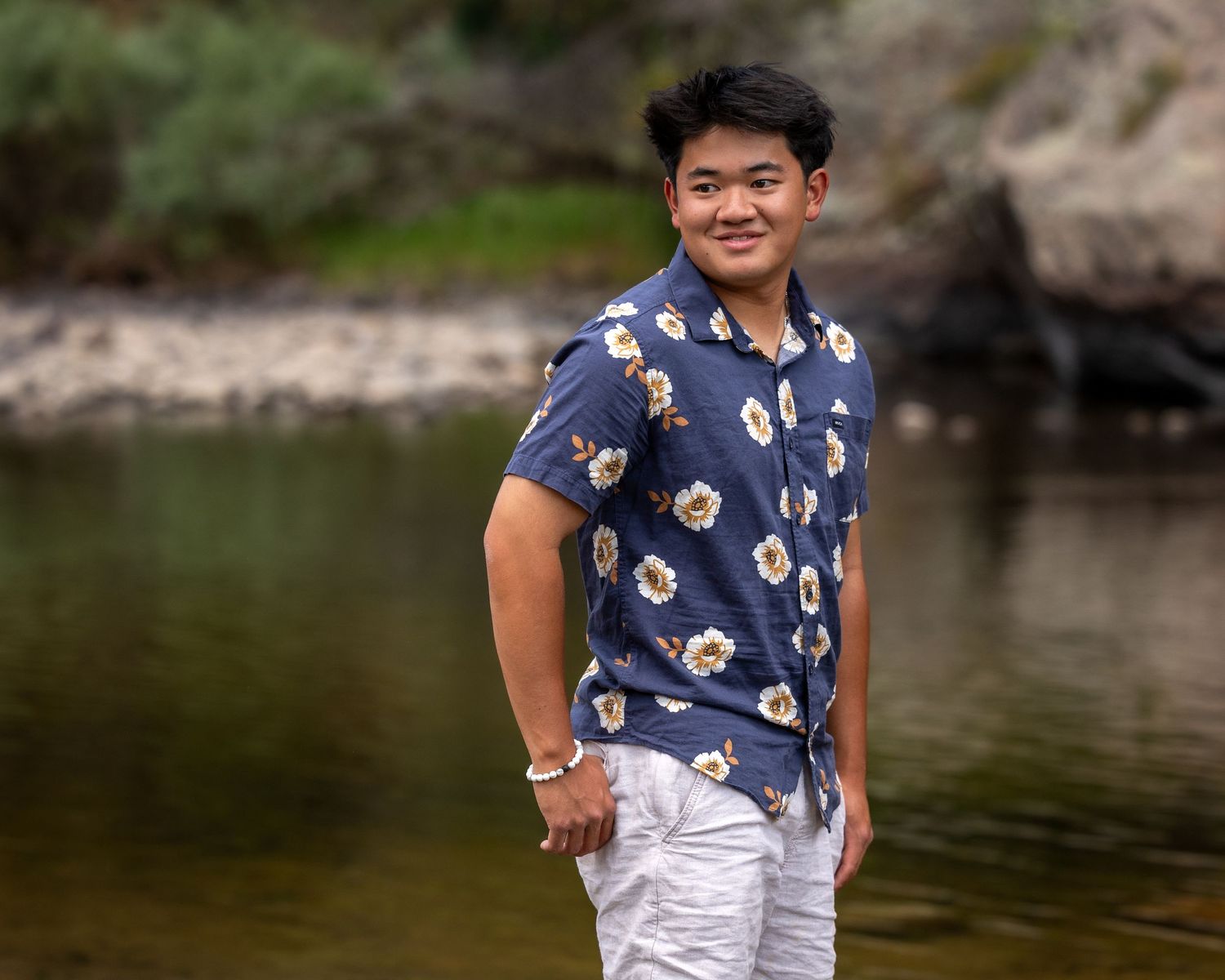Smiling young man in floral navy shirt standing by a rocky creek outdoors.