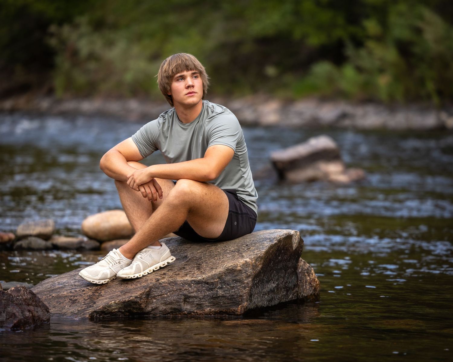 Teen boy in gray shirt sitting on river rocks with arms around knees outdoors.