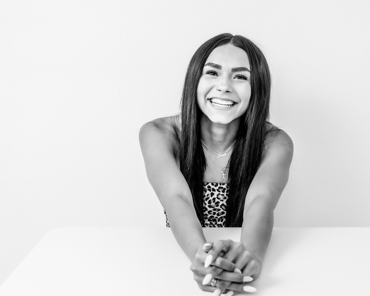 Black and white portrait of a smiling young woman leaning forward on a white surface in studio.