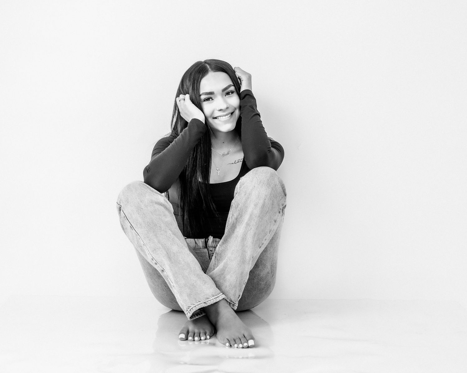 Young woman with dark hair sits cross-legged on the floor smiling in a black and white studio portrait.