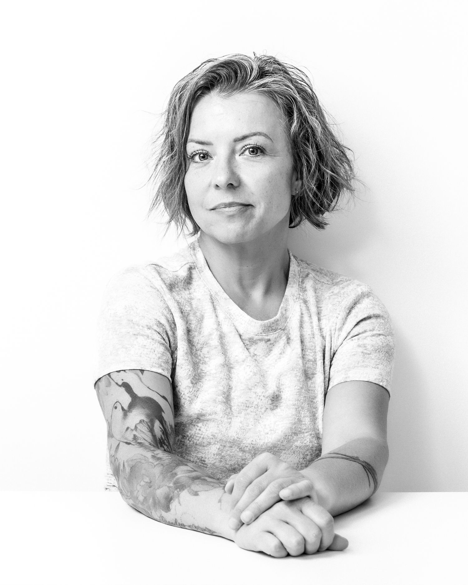 Black and white portrait of a woman with short hair and tattoos sitting at a table.