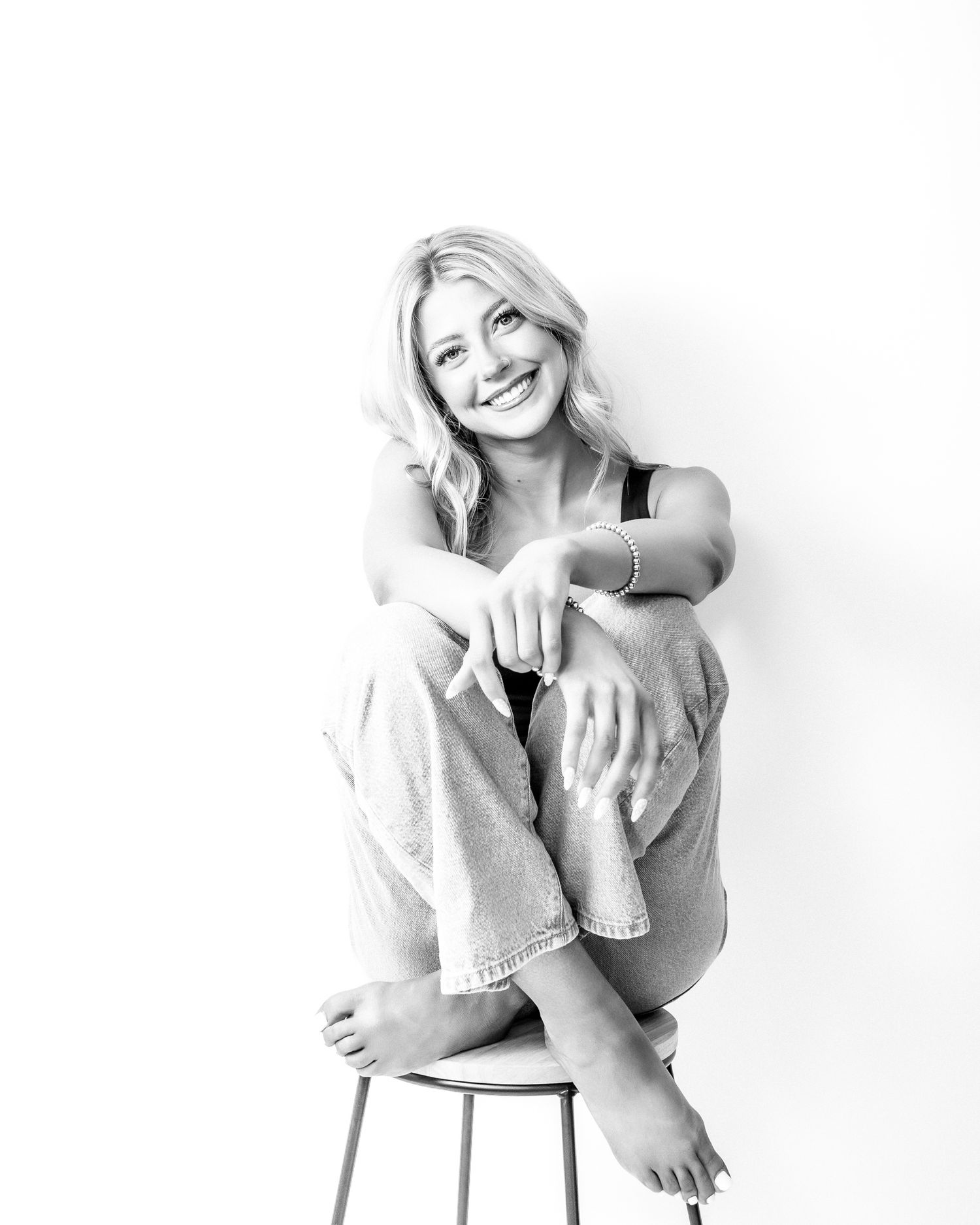 Smiling blonde woman sitting barefoot on a stool, posing casually in jeans, black and white photo.