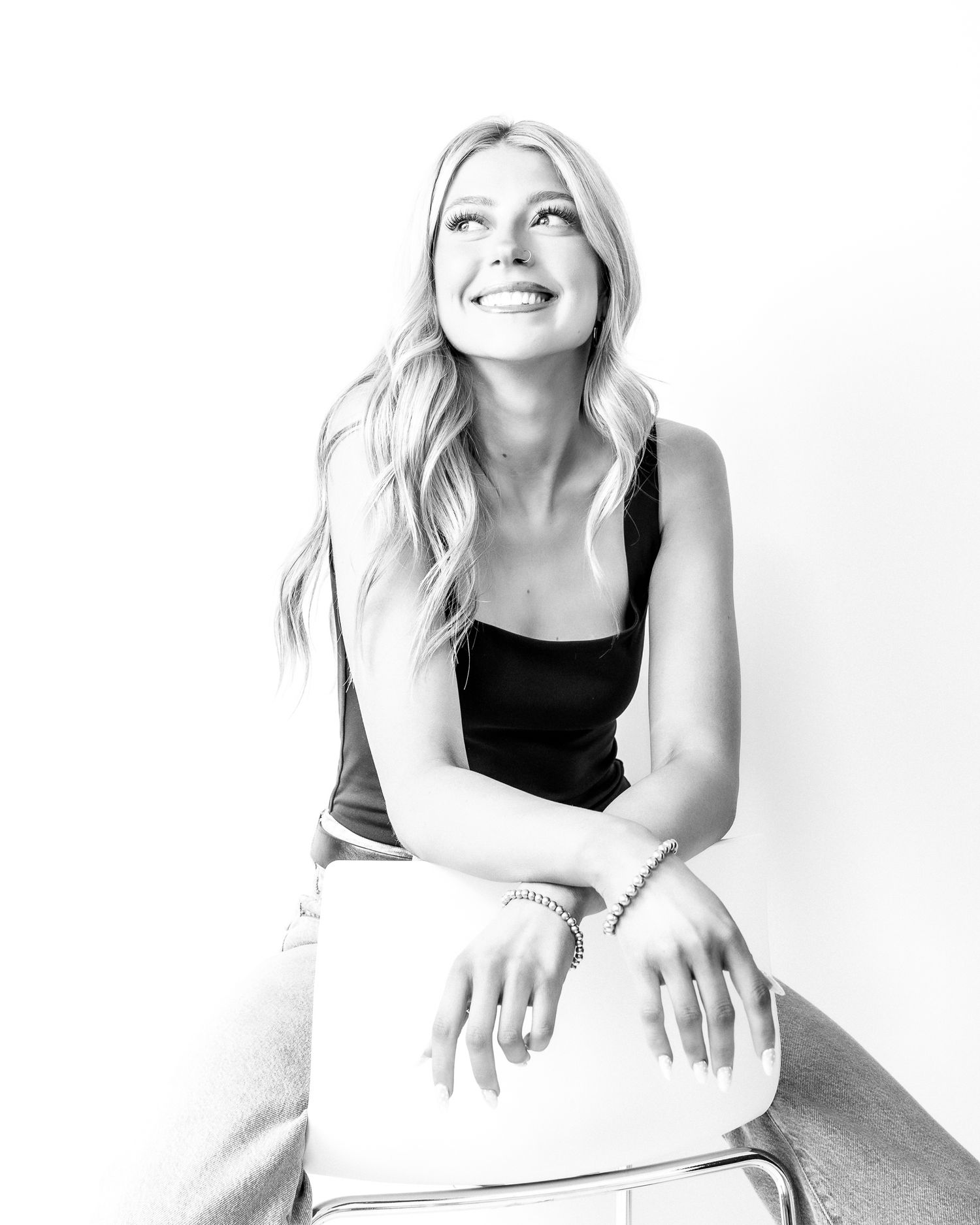 Joyful blonde woman in black tank top sitting on stool looking up in black and white studio photo.