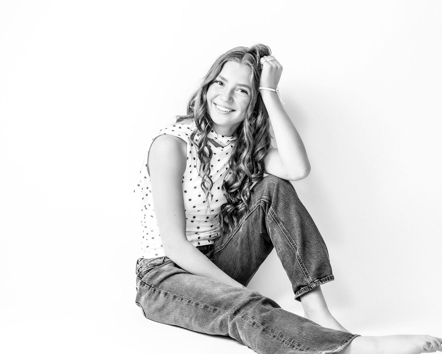 Young woman with wavy hair sits on the floor smiling in a polka dot top in a black and white portrait.