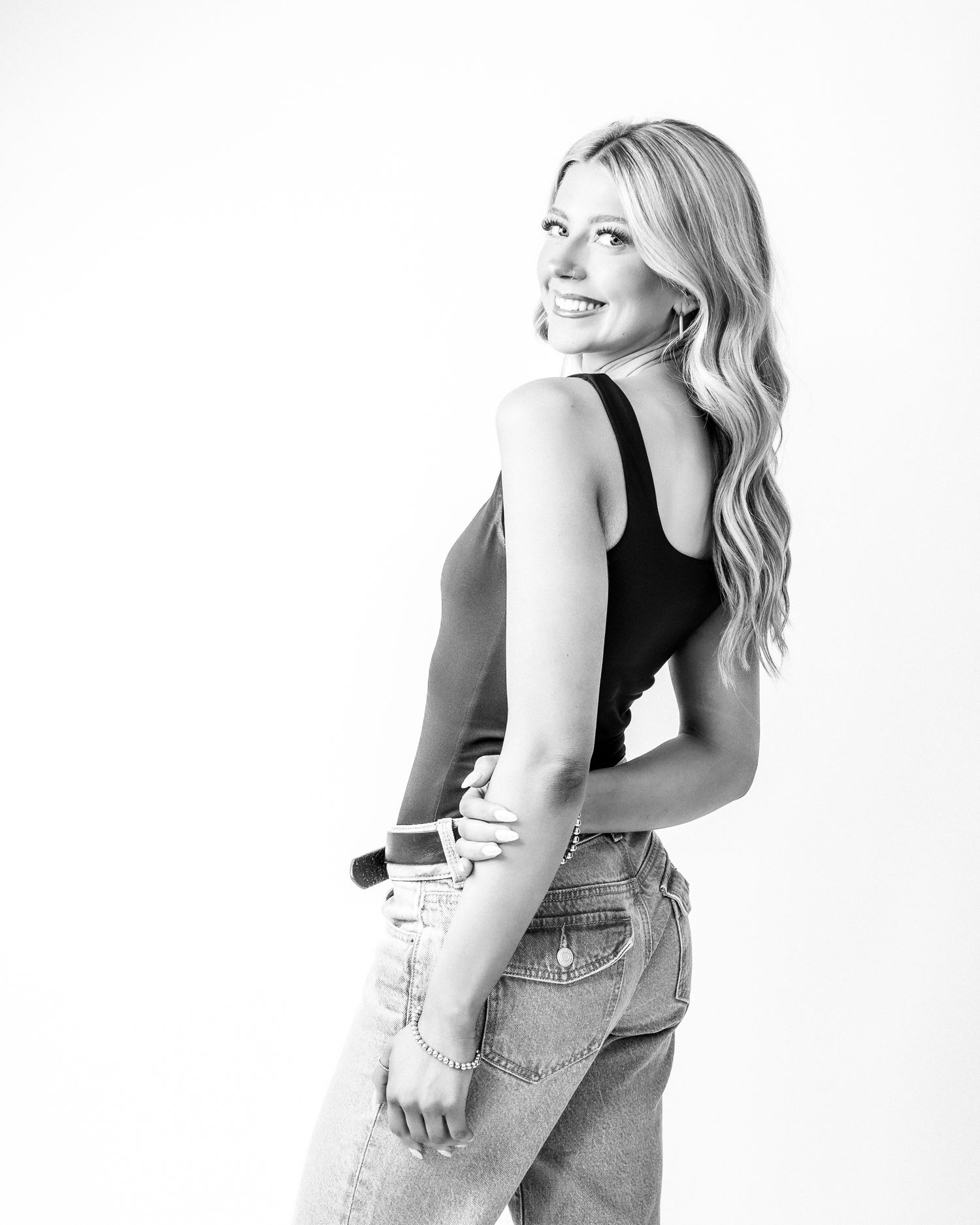 Black and white photo of a smiling blonde woman in jeans and tank top holding a makeup brush.