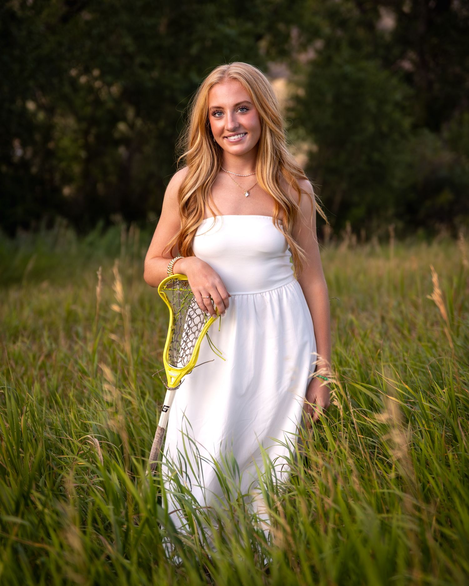 Blonde teen girl in white dress holds yellow lacrosse stick while standing in tall grass.