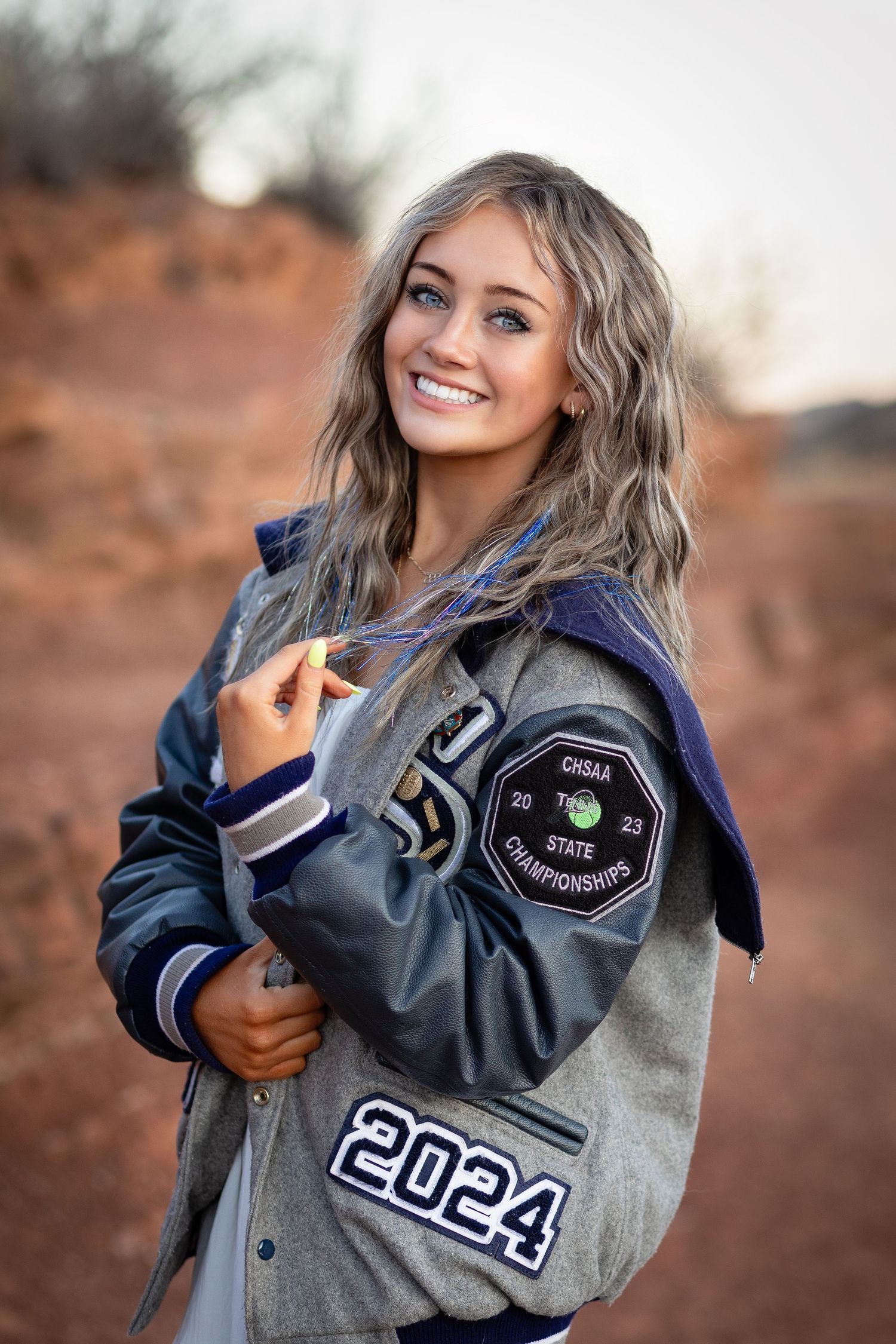 Smiling blonde girl in 2024 letterman jacket posing in front of red rock landscape.