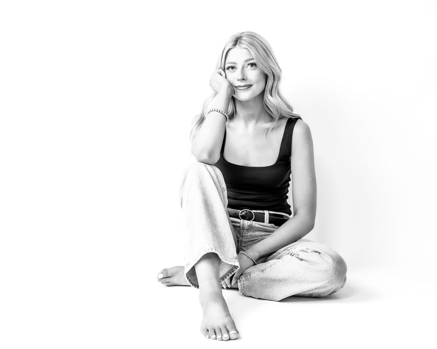 Black and white photo of a smiling blonde woman sitting cross-legged on a white background in casual attire.