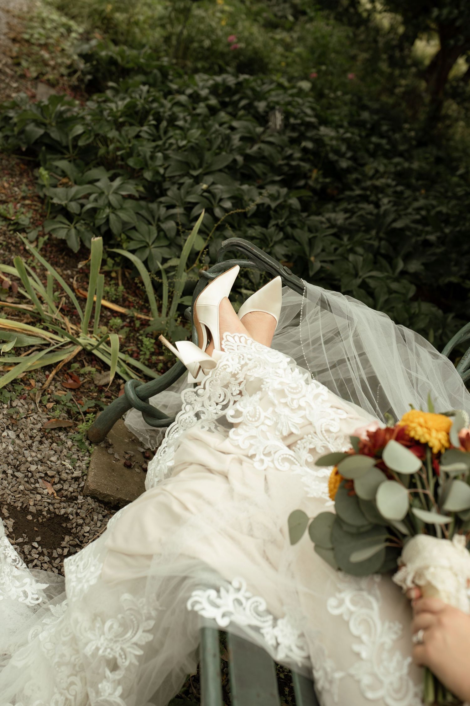 Close-up of bride in lace gown wearing white heels holding autumn bouquet while seated on garden bench.