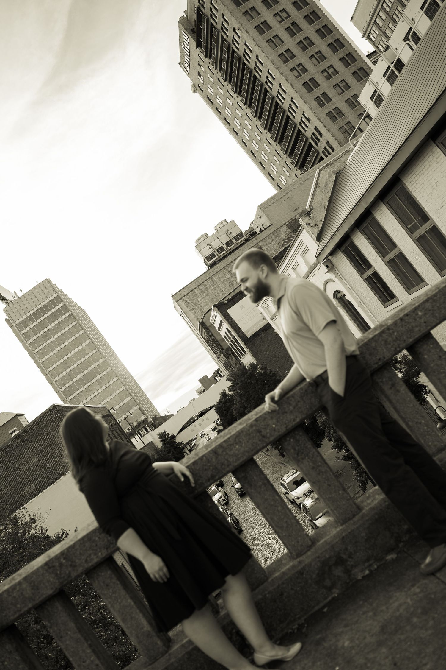 Black and white photo of a couple on urban steps with tall city buildings towering in the background.
