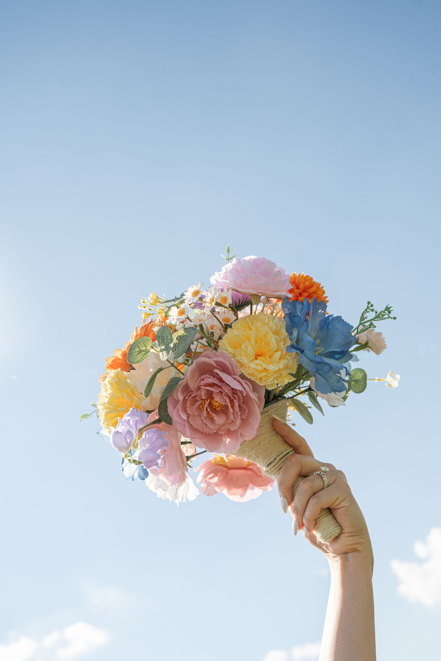 Hand raises colorful spring wedding bouquet of roses and mixed flowers against blue sky.
