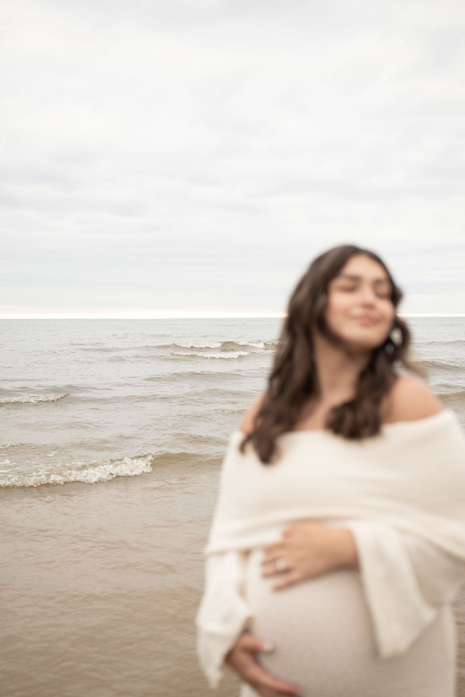 Pregnant woman in white wrap standing peacefully on a beach with ocean waves in the background.