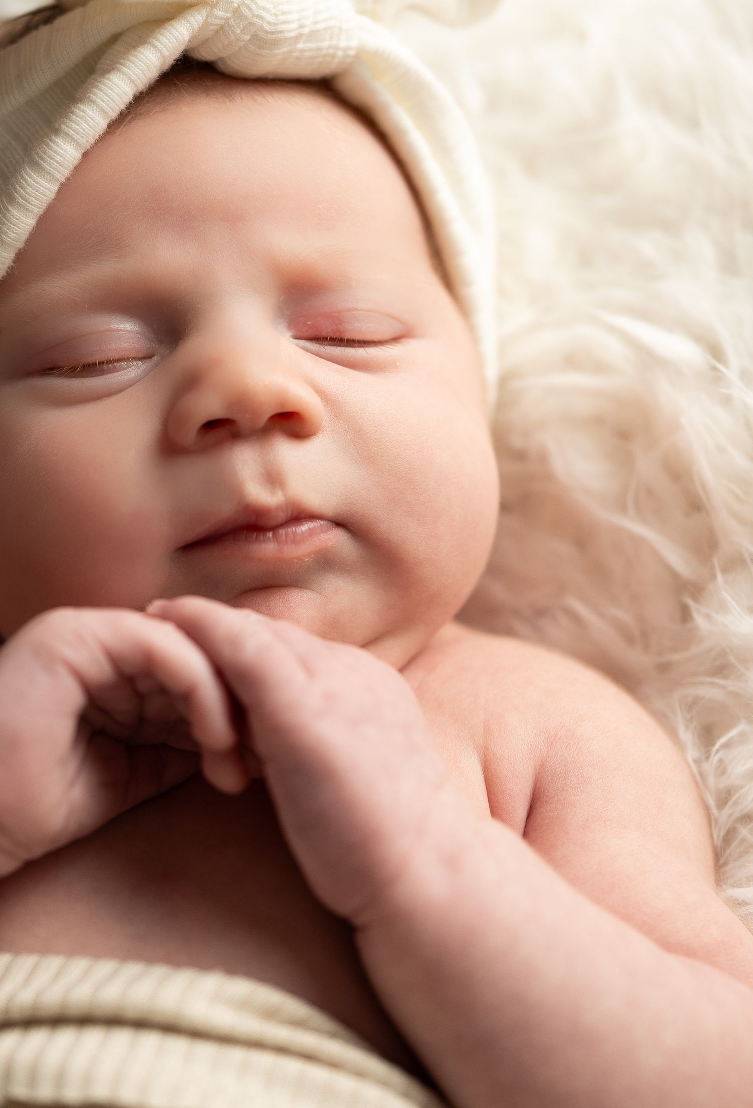 Sleeping newborn baby wearing a cream knit hat resting on a soft fluffy white blanket.