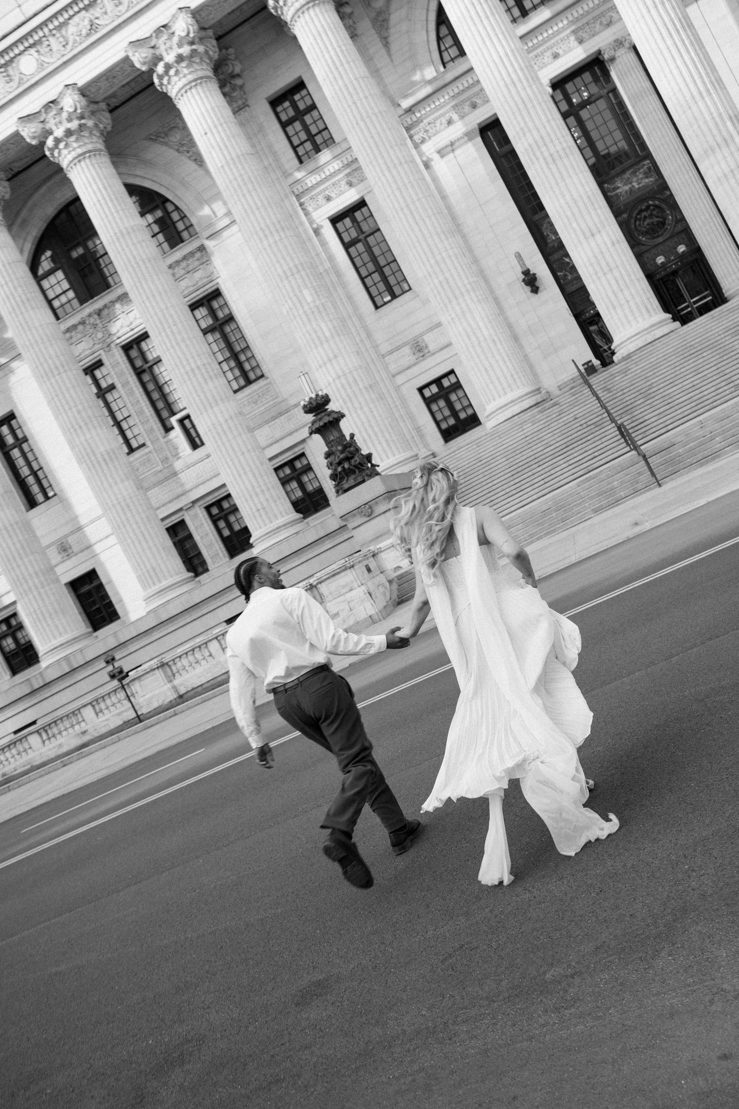 Black and white photo of a bride and groom running joyfully in front of a grand columned building.