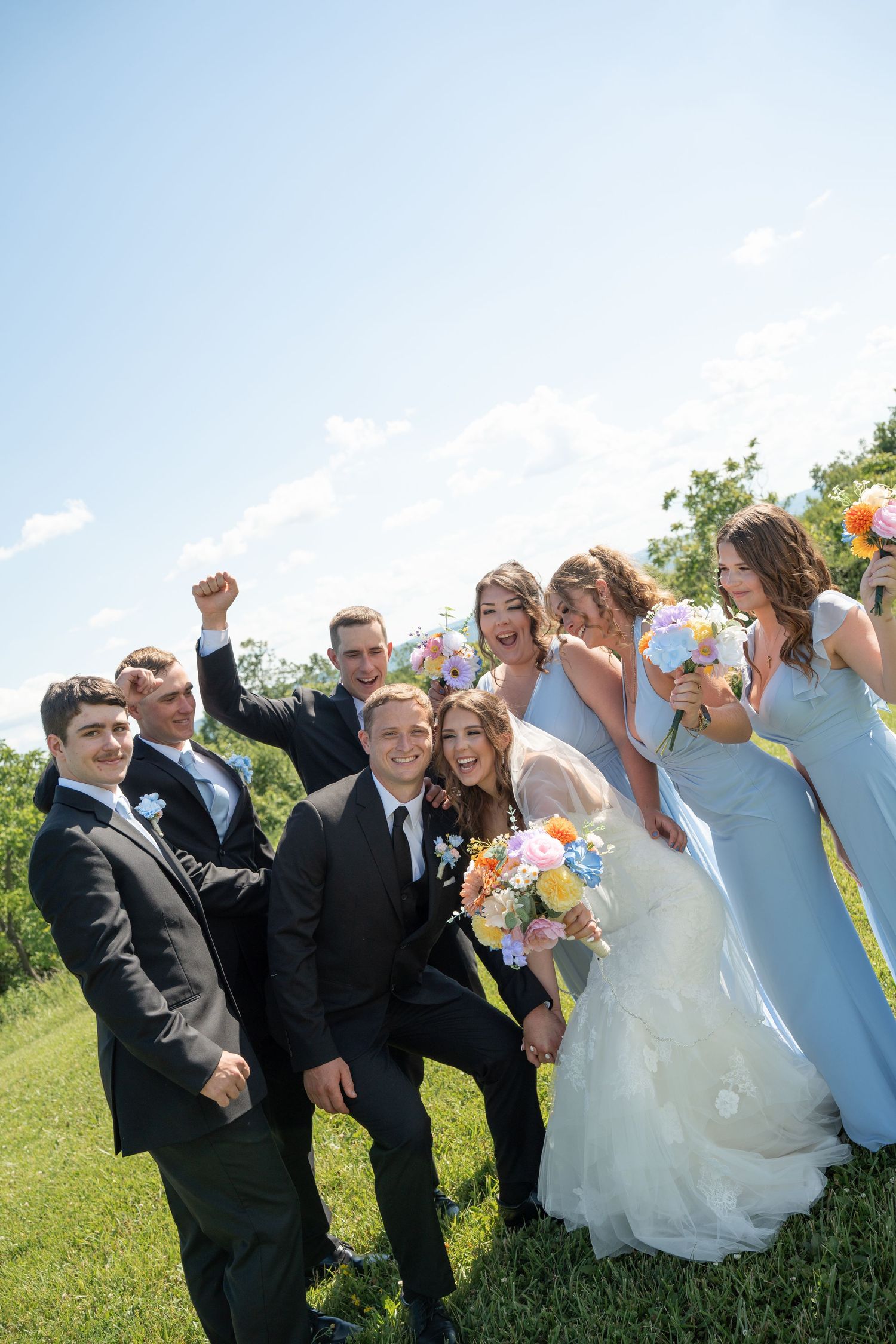Wedding party celebrating outdoors on sunny day, bride holding colorful bouquet surrounded by bridesmaids in blue and groomsmen.