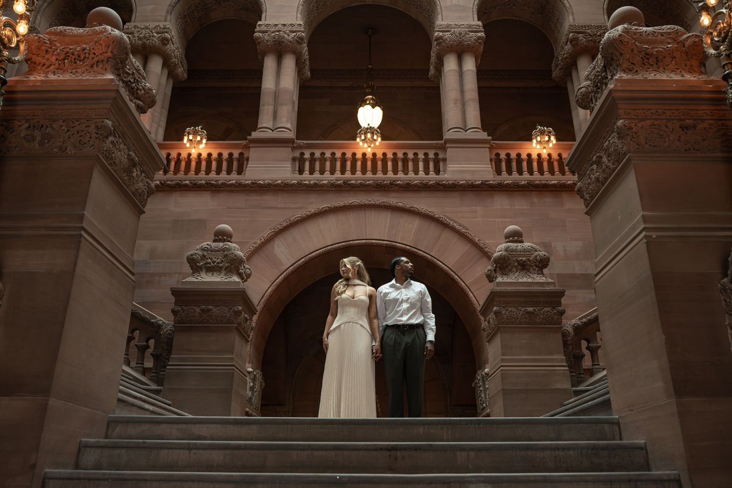 Bride and groom standing together on grand staircase inside ornate historic building with arched stone architecture.