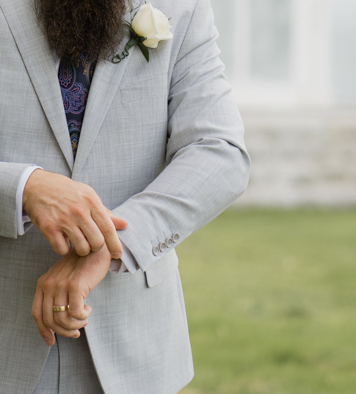 Groom in light gray suit adjusting cufflinks with white rose boutonniere and gold wedding band visible.