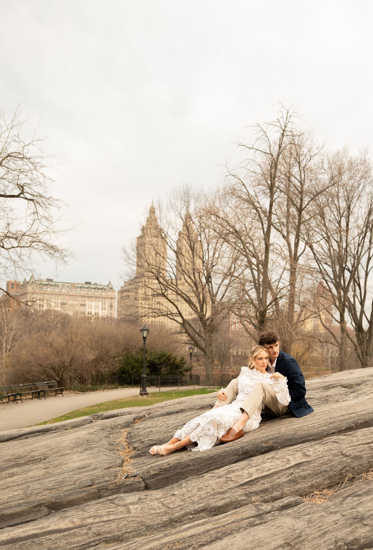 Couple sitting on rocks in Central Park, NYC, sharing a romantic moment with bare trees and buildings in the background.