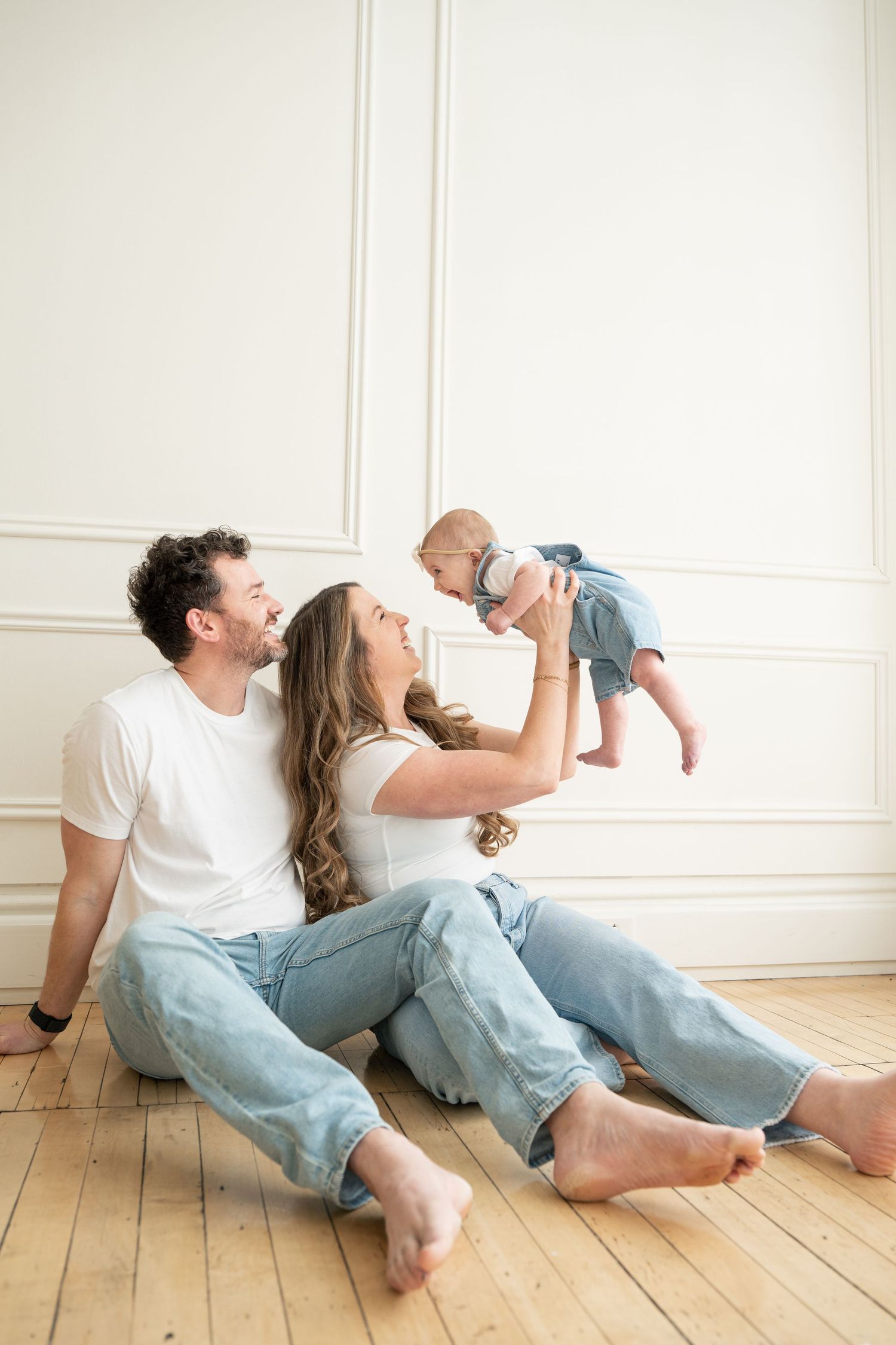 Happy couple sits on hardwood floor lifting their baby in a bright white room.