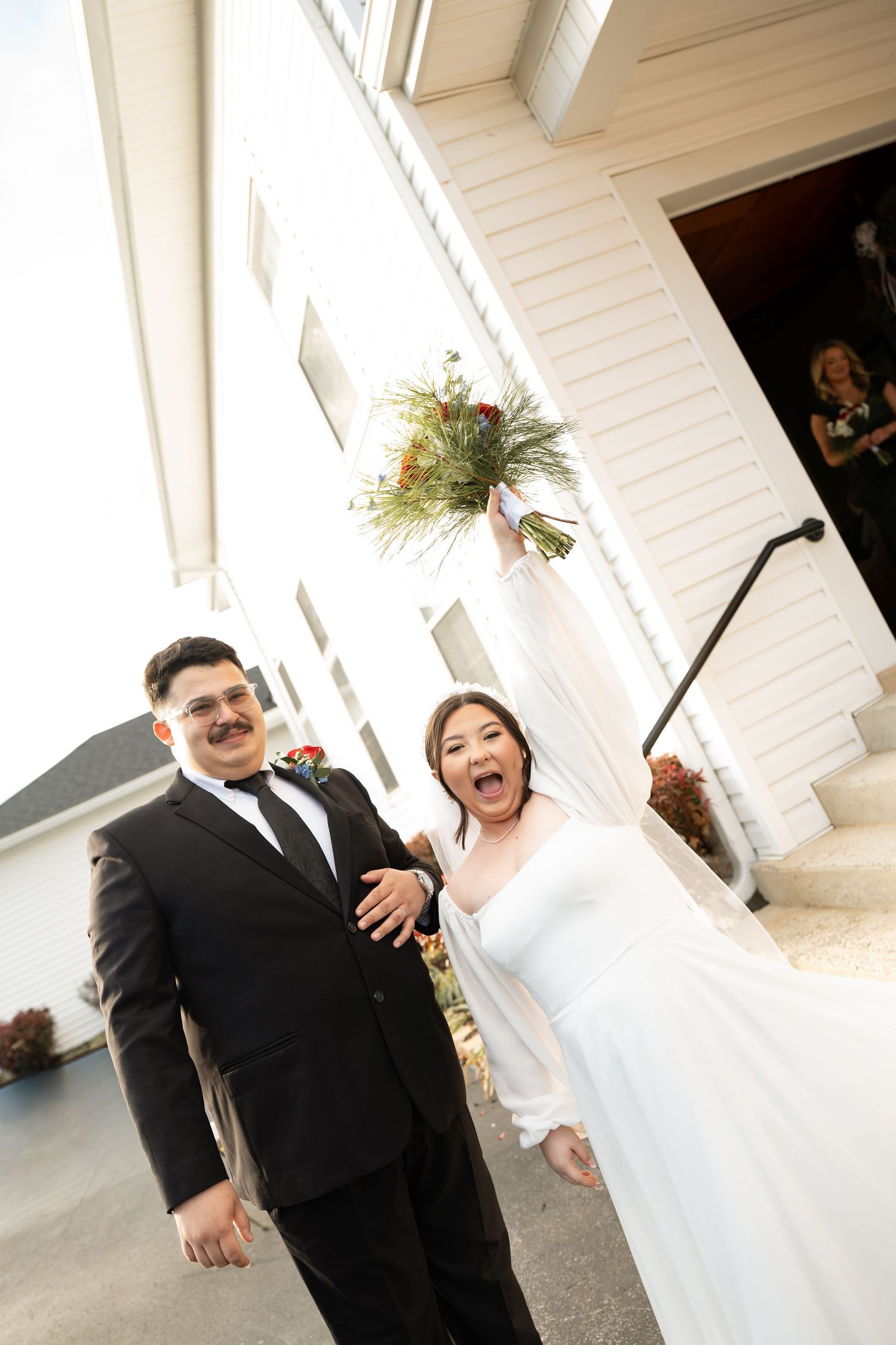 Excited bride and groom celebrate outside white chapel holding colorful wedding bouquet.